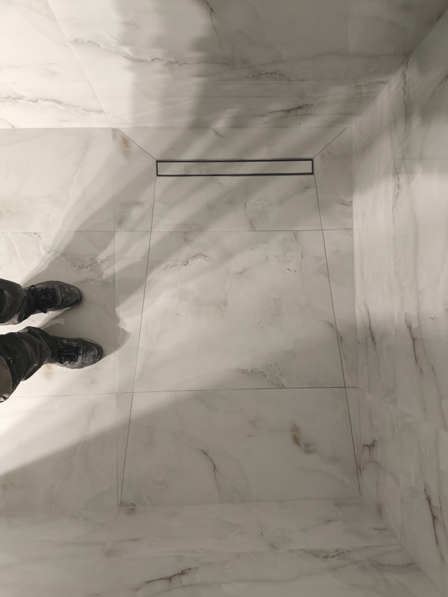 Ukończona posadzka z białych, marmurkowych płytek w kabinie prysznicowej z odpływem liniowym. Widoczne stopy w roboczych butach.