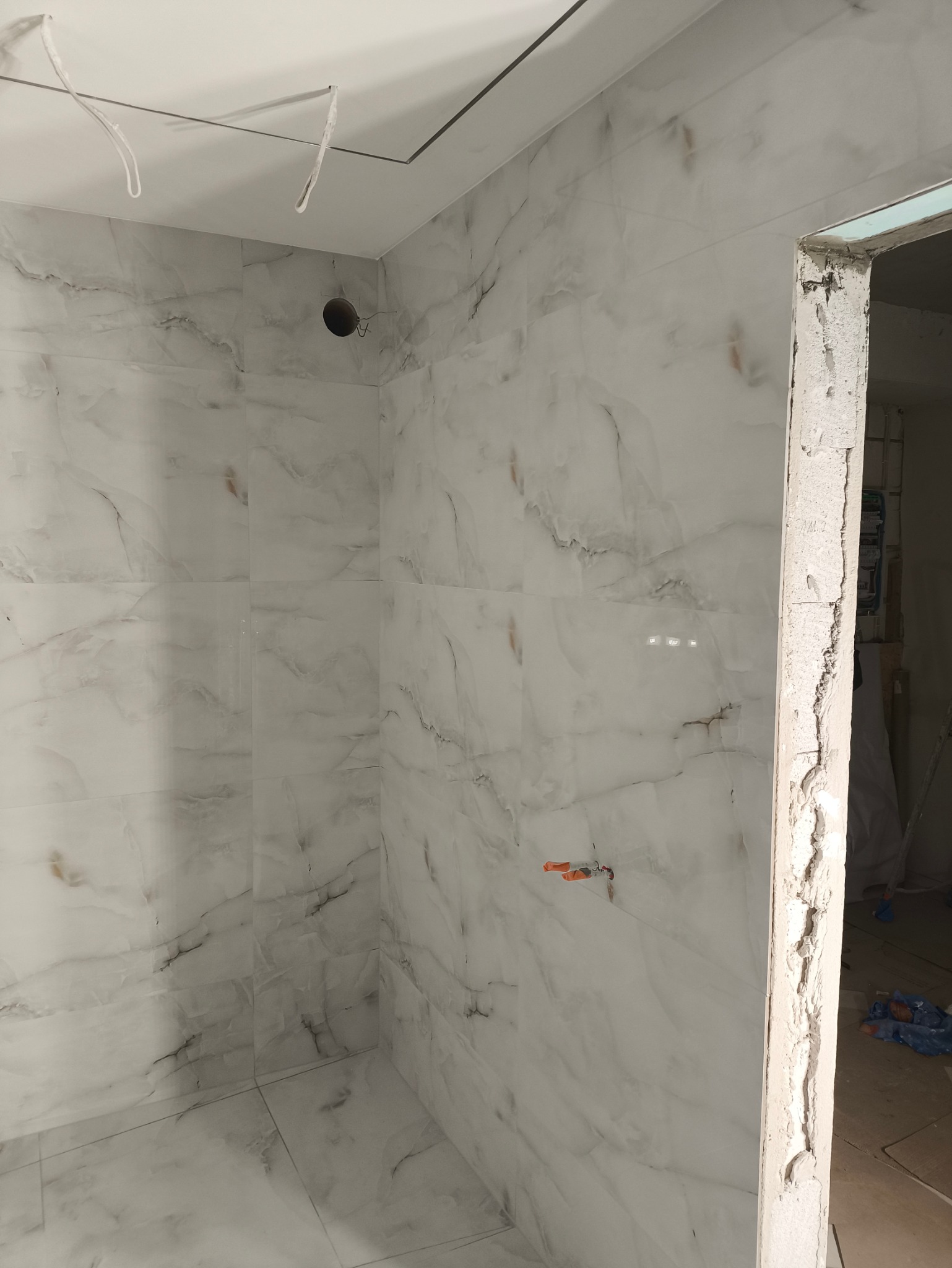 Ściany i podłoga wyłożone białymi płytkami imitującymi marmur w nowej łazience, widoczne kable wystające z sufitu i surowa ościeżnica drzwi.