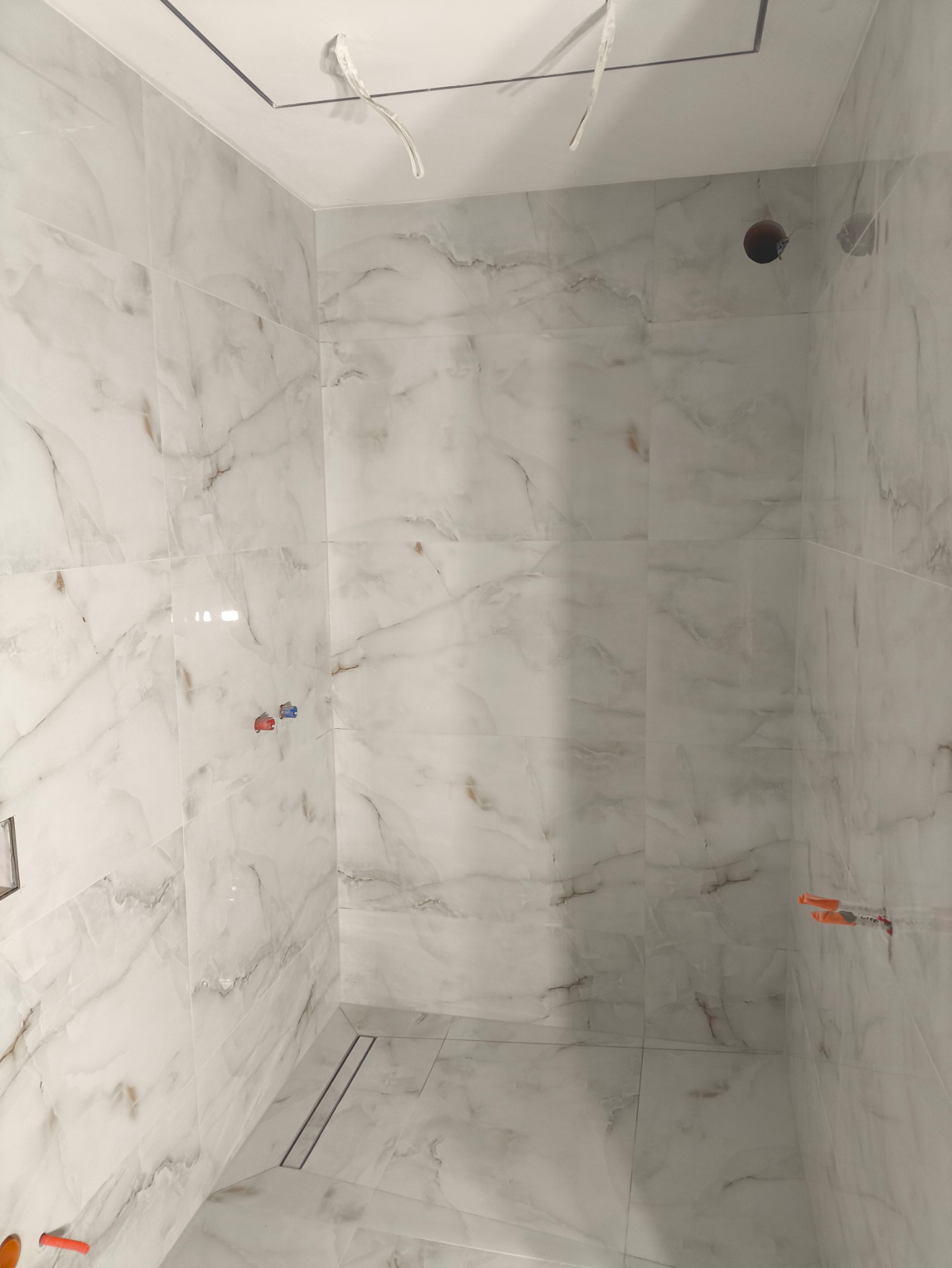 Wykończona kabina prysznicowa z białymi, marmurkowymi płytkami, widoczne wystające kable ze ściany i sufitu, oraz odpływ liniowy w podłodze.