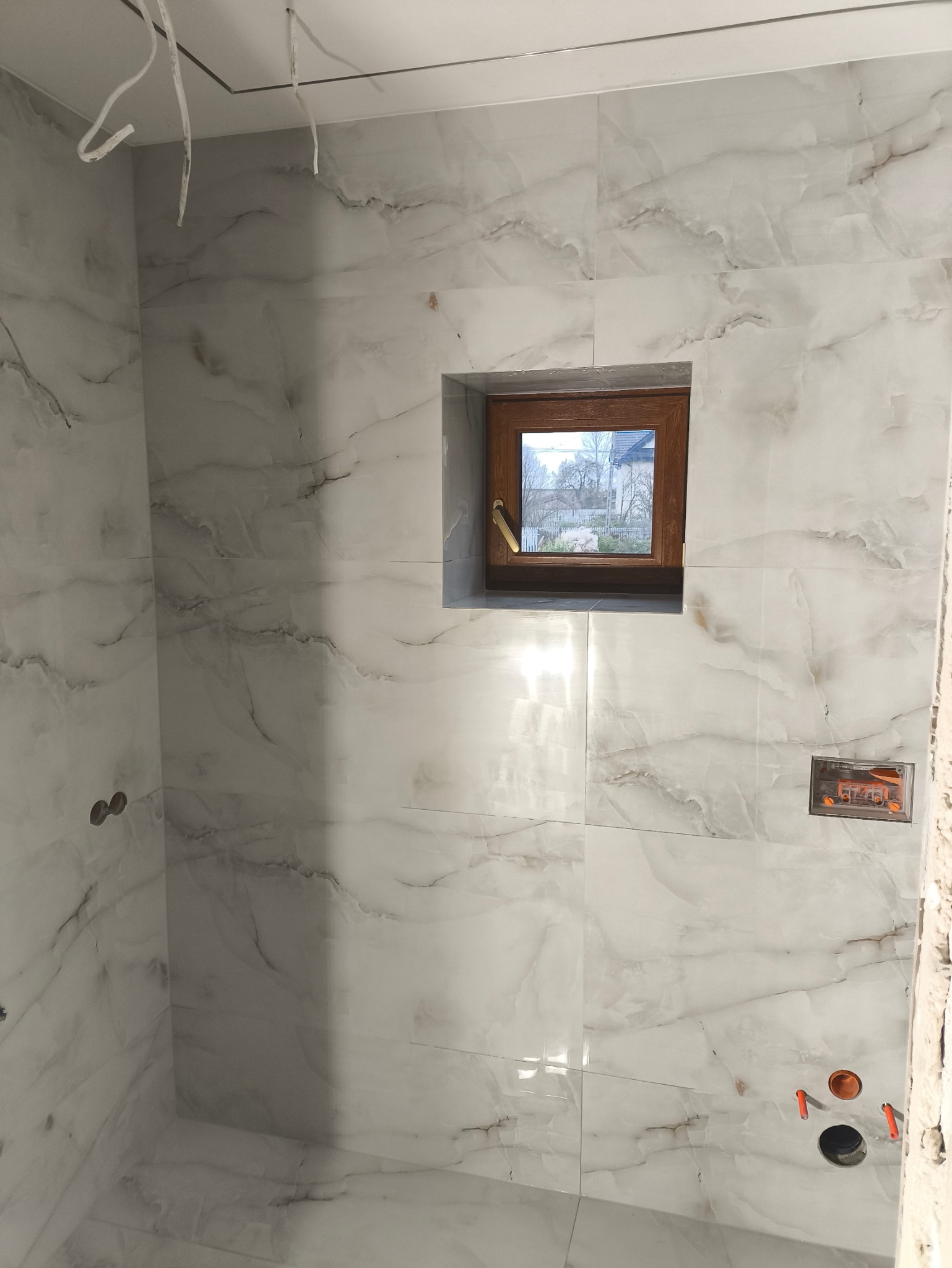 Ściana łazienki wyłożona białymi, marmurkowymi płytkami z małym drewnianym oknem i wystającymi przewodami oraz puszkami instalacyjnymi w trakcie prac wykończeniowych.