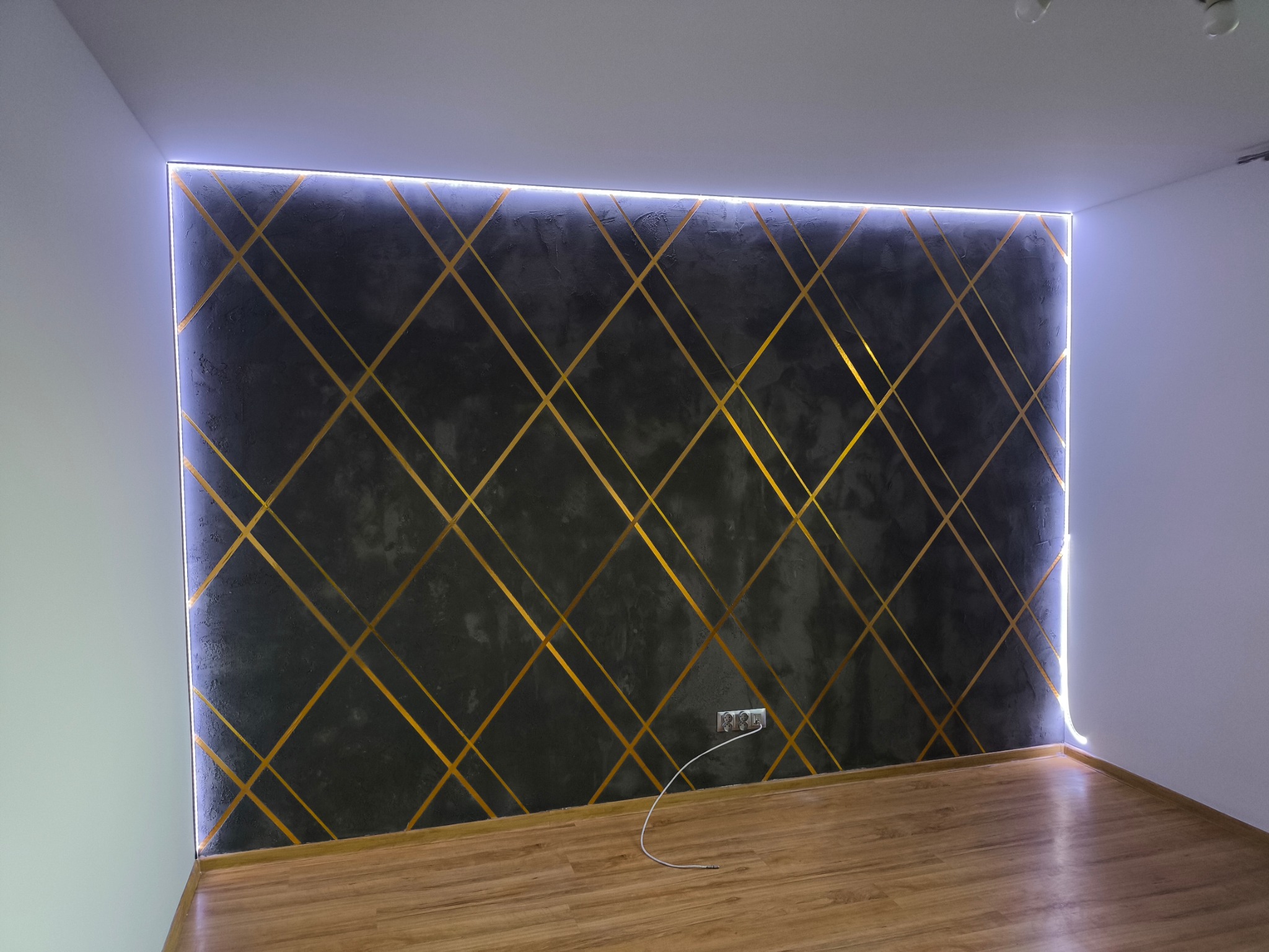 Ściana z dekoracyjnym tynkiem w odcieniach szarości, z geometrycznym wzorem ze złotych pasków i podświetleniem LED w Radomiu.