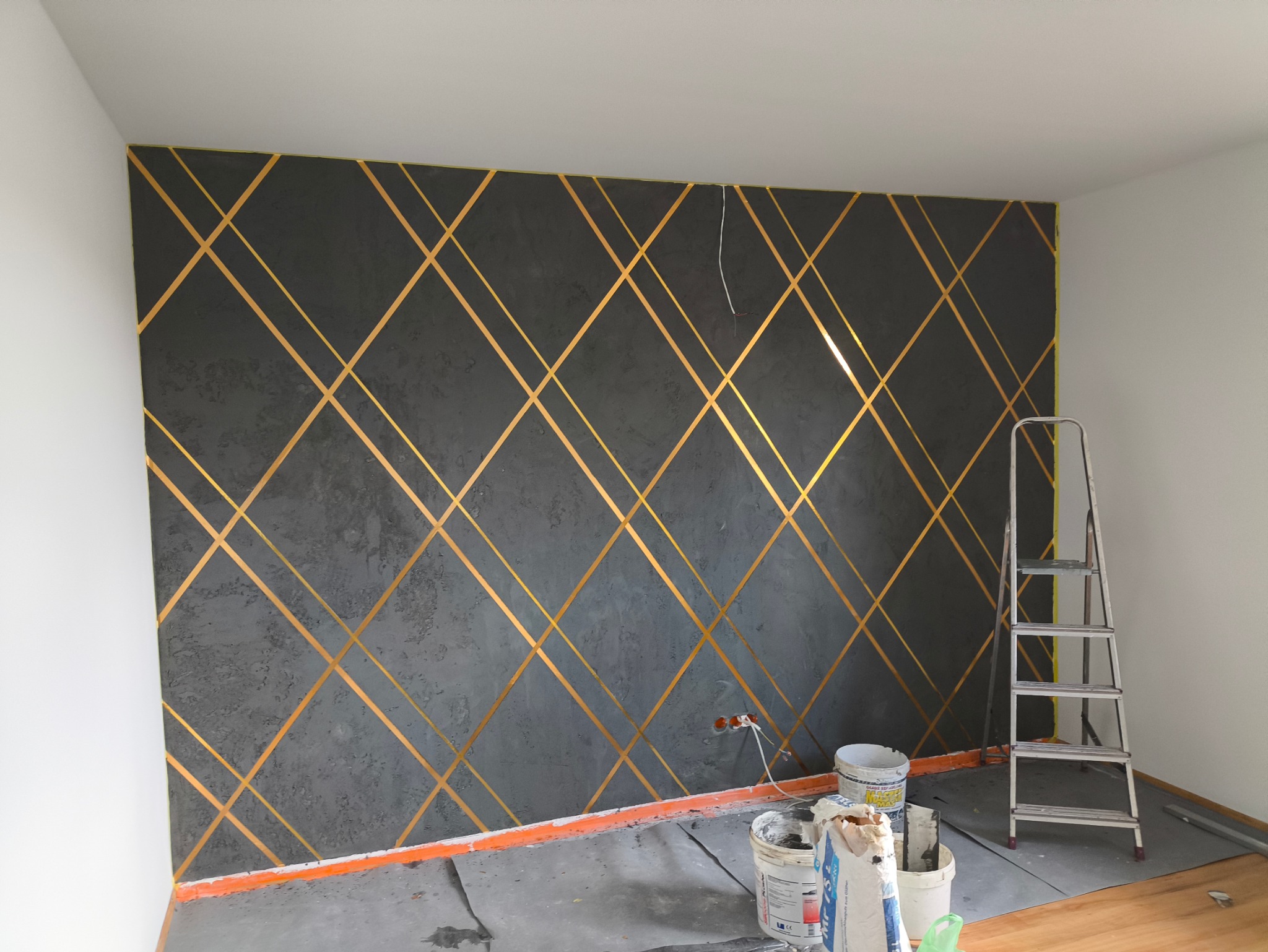 Ściana pomalowana na szaro z geometrycznym wzorem z taśm w kolorze złotym, drabina oparta o ścianę, widoczne puszki z farbą i narzędzia malarskie na podłodze.