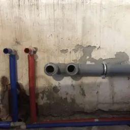 Surowy stan ściany z widocznymi, nowo zamontowanymi rurami instalacji wodnej i kanalizacyjnej: niebieskie i czerwone rury z tworzywa, szara rura kanalizacyjna wchodząca w ścianę.