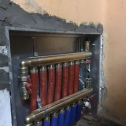 Rozdzielacz ogrzewania podłogowego z czerwonymi i niebieskimi rurami zamontowany w ścianie z widocznymi śladami po remoncie.