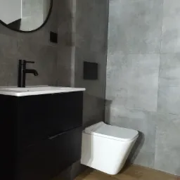 Nowoczesna łazienka z szarymi płytkami, drewnopodobną podłogą, czarną szafką umywalkową z minimalistyczną baterią i podwieszaną toaletą.