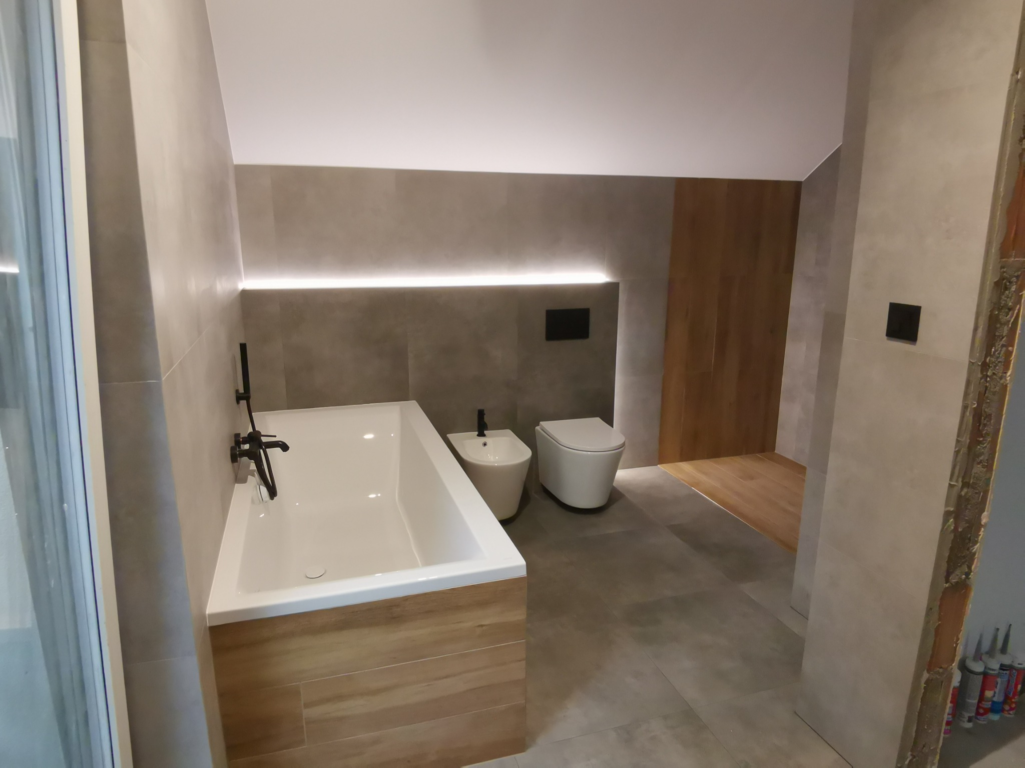 Nowoczesna łazienka z wanną obudowaną drewnem, bidetem i toaletą w minimalistycznym stylu, oświetlenie LED pod sufitem i szare płytki na ścianach i podłodze, widoczne materiały budowlane przy...