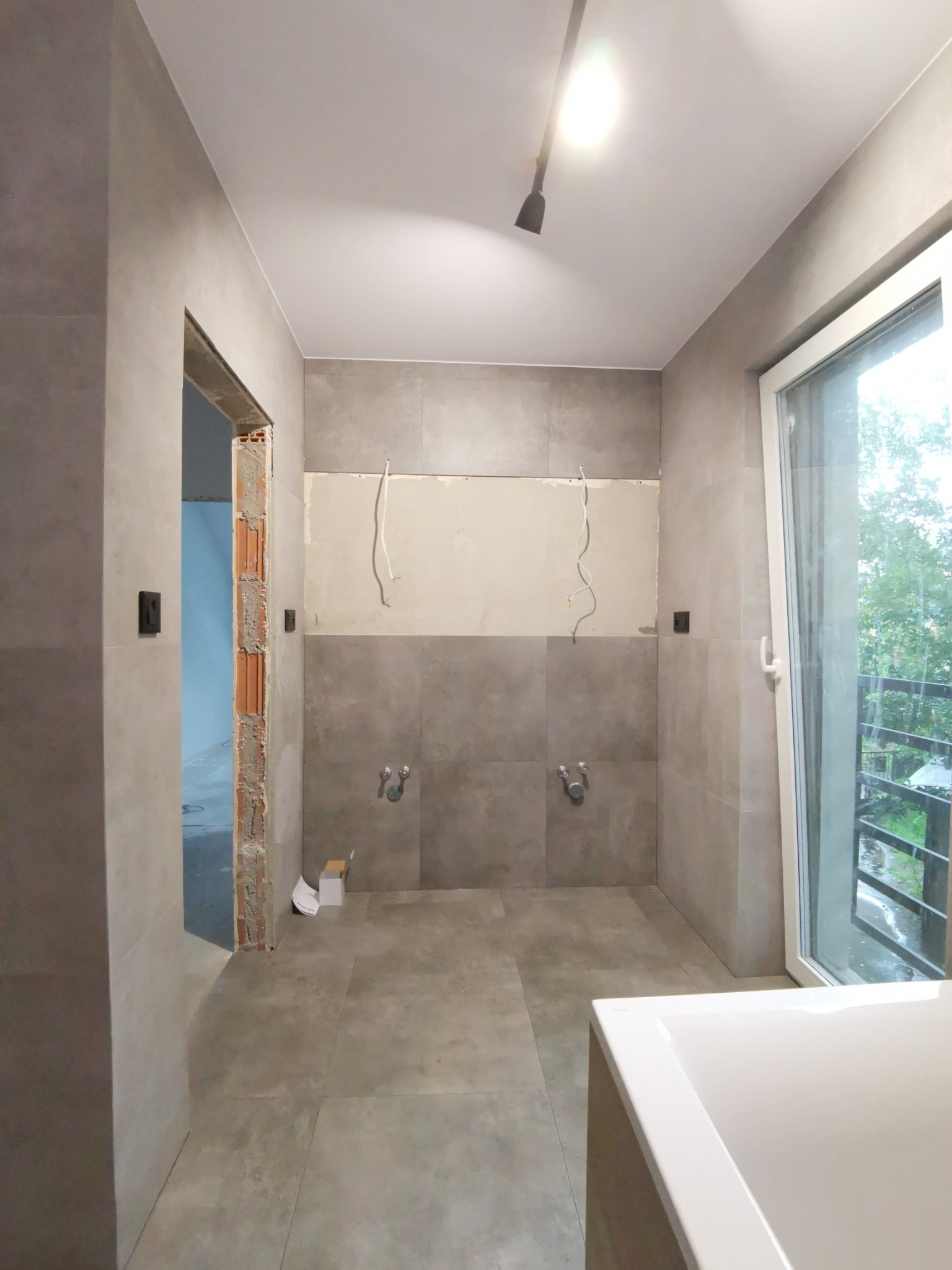 Łazienka w trakcie prac wykończeniowych: szare płytki na ścianach i podłodze, przygotowane przyłącza wodne, widoczne przewody elektryczne, surowa ościeżnica drzwiowa, okno z widokiem na zieleń.