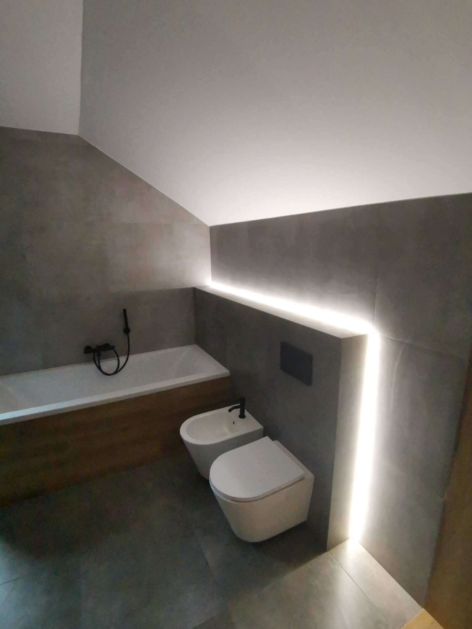 Nowoczesna łazienka w szarościach z podświetleniem LED, wisząca toaleta i bidet, bateria wannowa w kolorze czarnym.