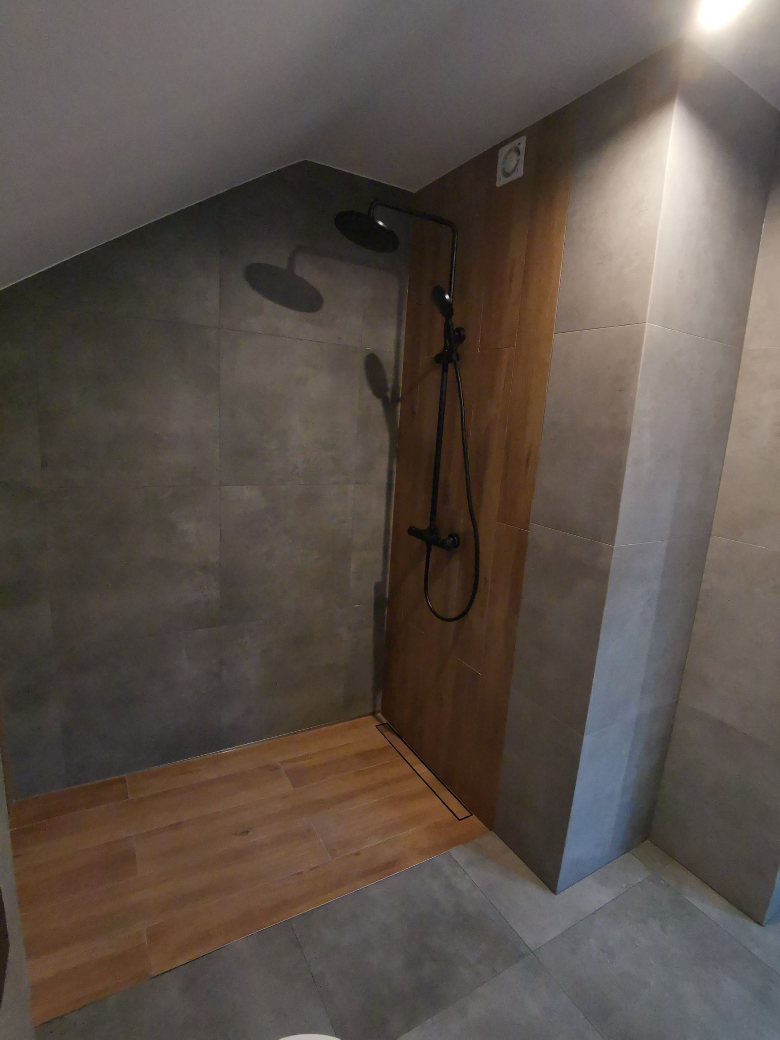 Nowoczesna kabina prysznicowa z deszczownicą w łazience na poddaszu, wykończona szarymi płytkami i drewnopodobną podłogą.