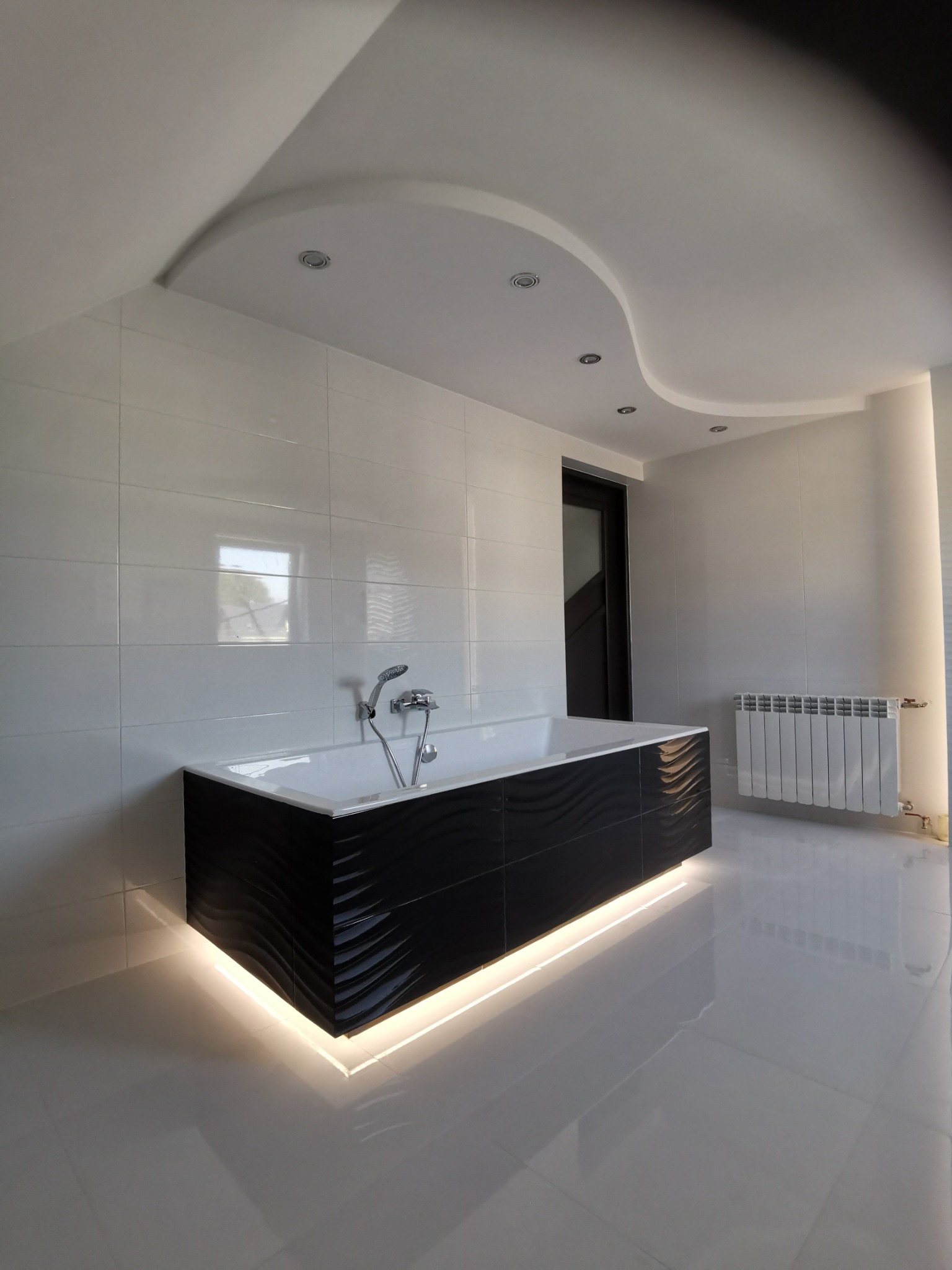Nowoczesna łazienka z białą ceramiką, czarną wanną z podświetleniem LED, białymi płytkami ściennymi i podłogowymi oraz dekoracyjnym podwieszanym sufitem z oświetleniem punktowym.