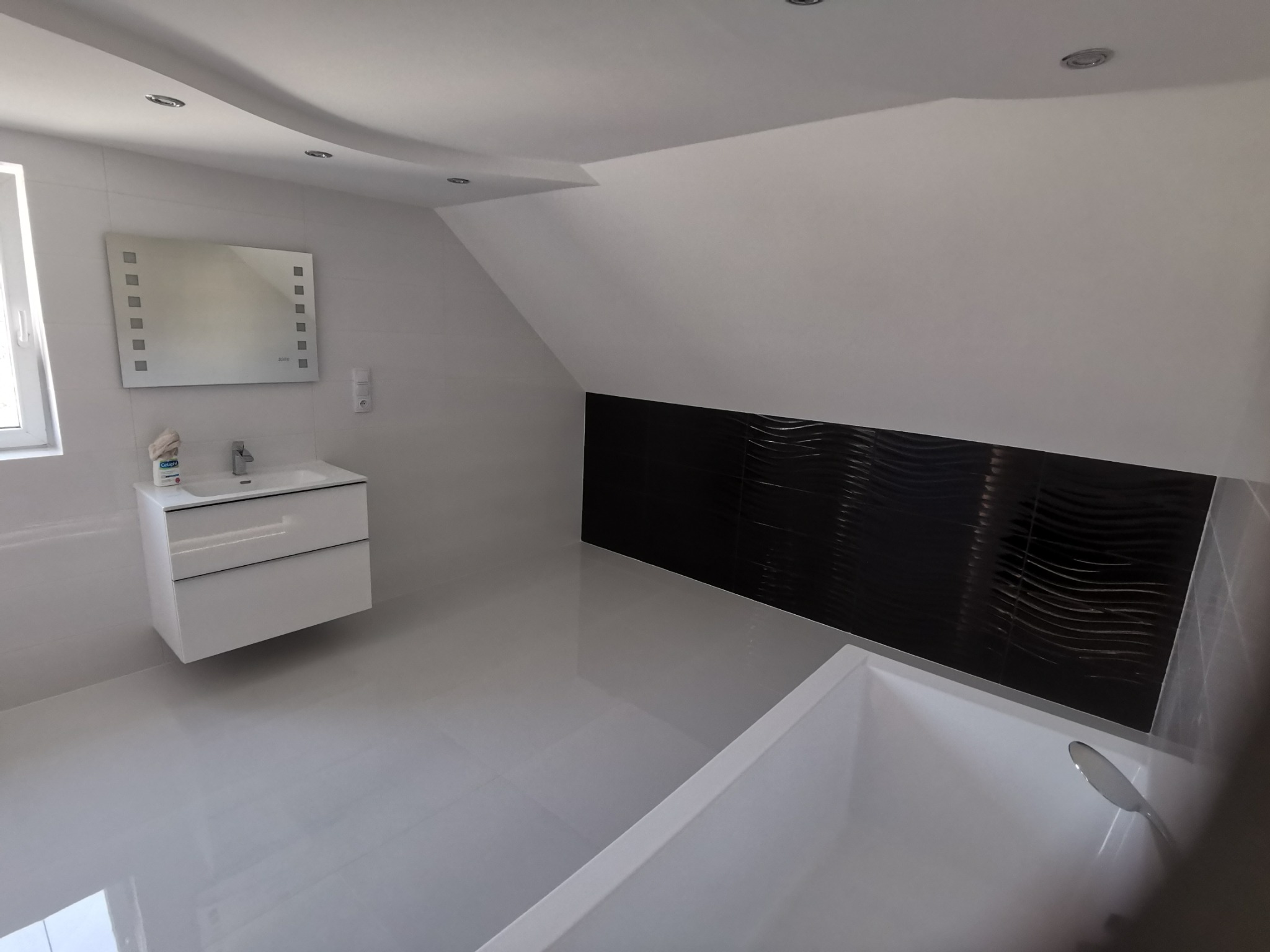 Nowoczesna łazienka na poddaszu z białą armaturą, czarną, falistą ścianą akcentującą i oświetleniem punktowym.