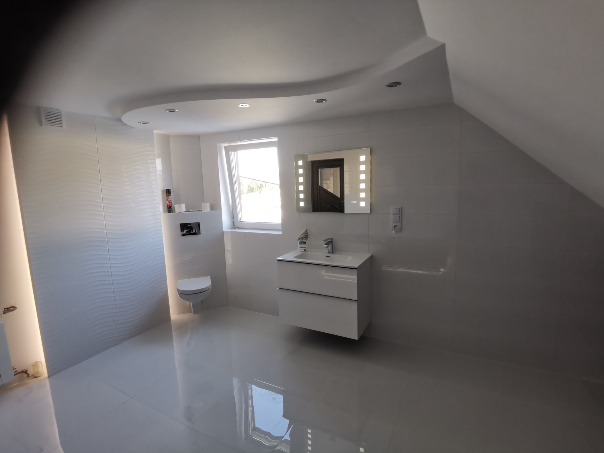 Nowoczesna łazienka na poddaszu z białymi płytkami o falistej strukturze, podświetlanym lustrem nad wiszącą szafką umywalkową i podwieszaną toaletą.