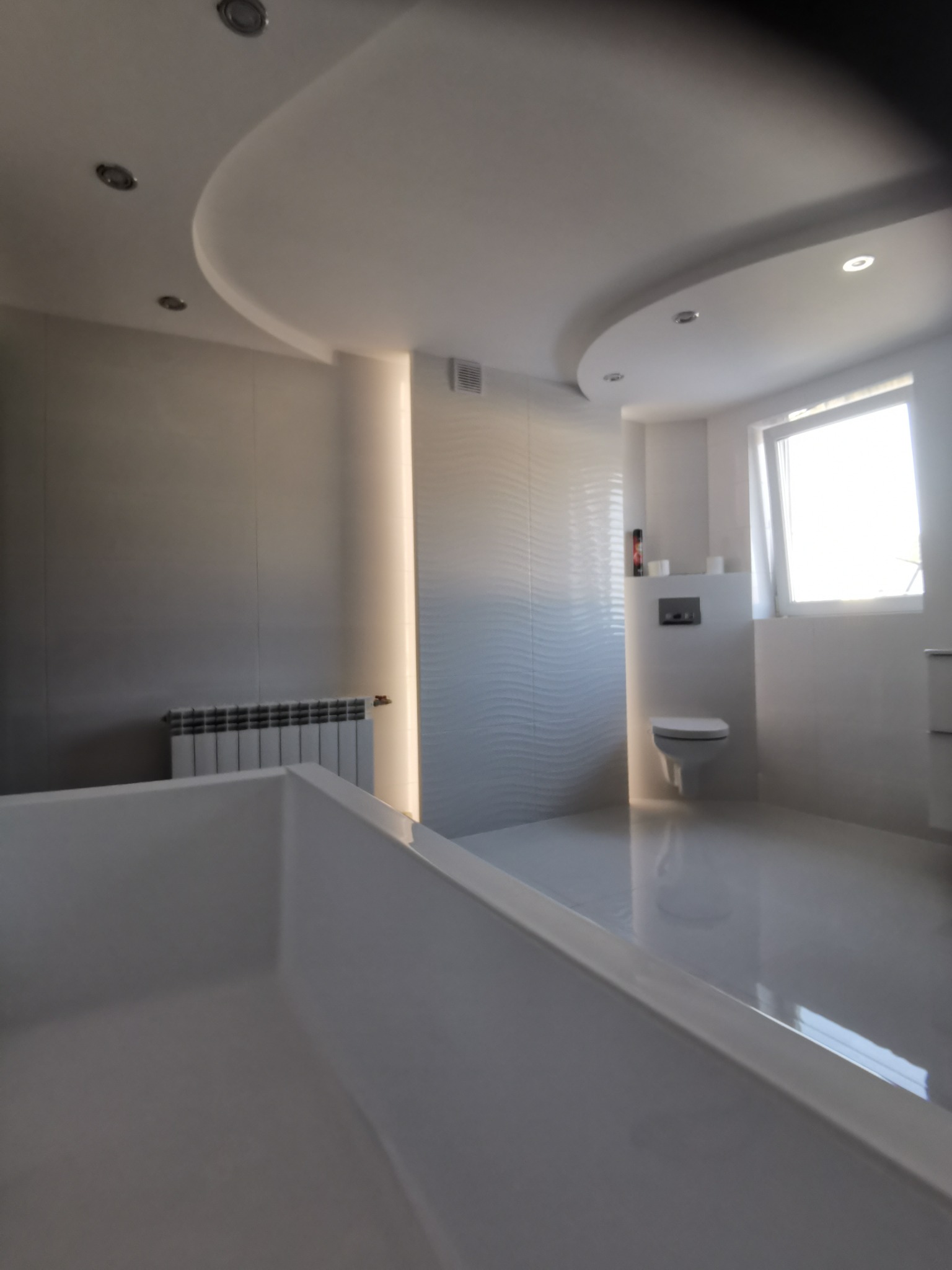 Nowoczesna łazienka z białą armaturą, podwieszaną toaletą, grzejnikiem żeberkowym i podświetlanym sufitem podwieszanym w kształcie łuku.