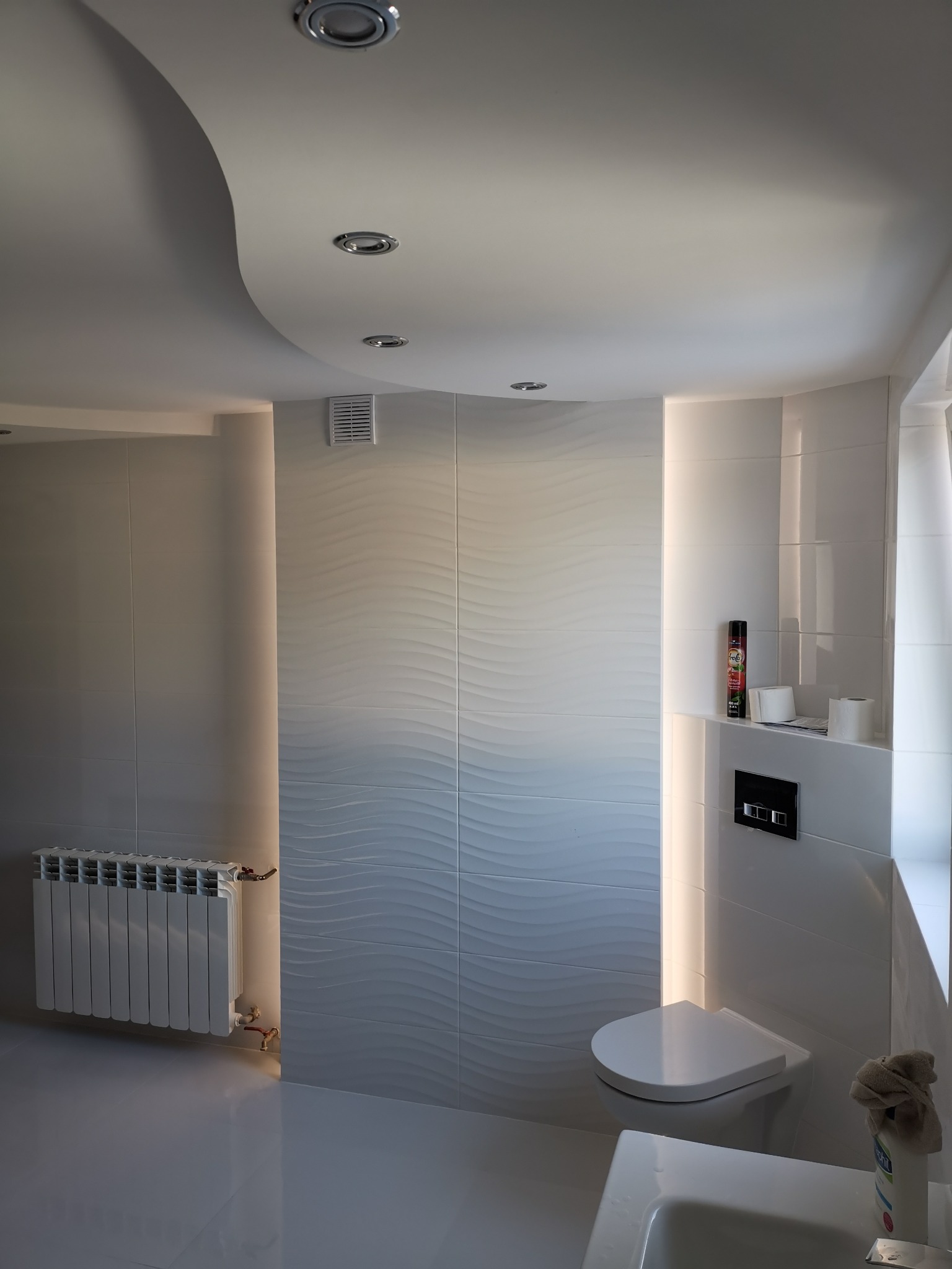 Nowoczesna łazienka wykończona białymi płytkami z falistym wzorem, podświetleniem LED, wiszącą toaletą i grzejnikiem żeberkowym.