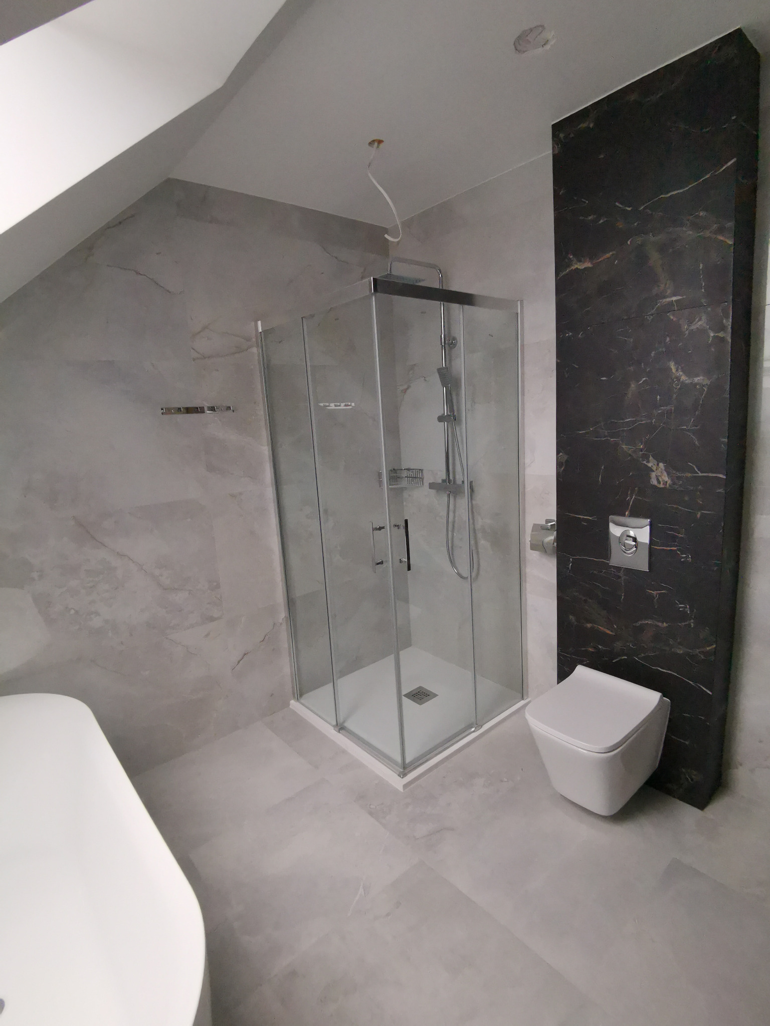 Nowoczesna łazienka na poddaszu z kabiną prysznicową, wiszącą toaletą i akcentem z czarnego marmuru na ścianie obok, całość utrzymana w szarościach.