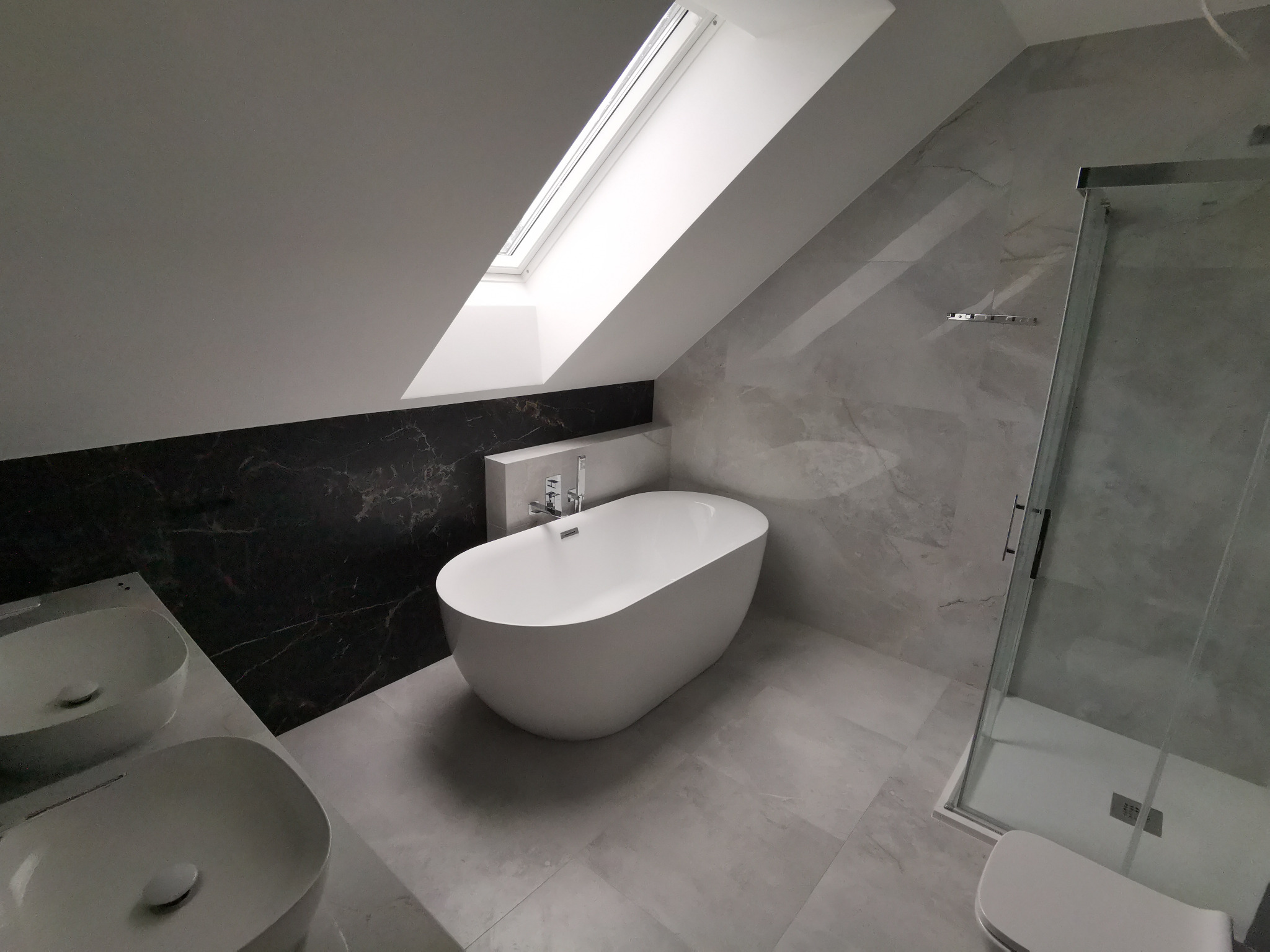 Nowoczesna łazienka na poddaszu z oknem dachowym, białą wolnostojącą wanną, umywalkami nablatowymi i kabiną prysznicową ze szklanymi drzwiami, wykończona płytkami imitującymi marmur w odcieniach...