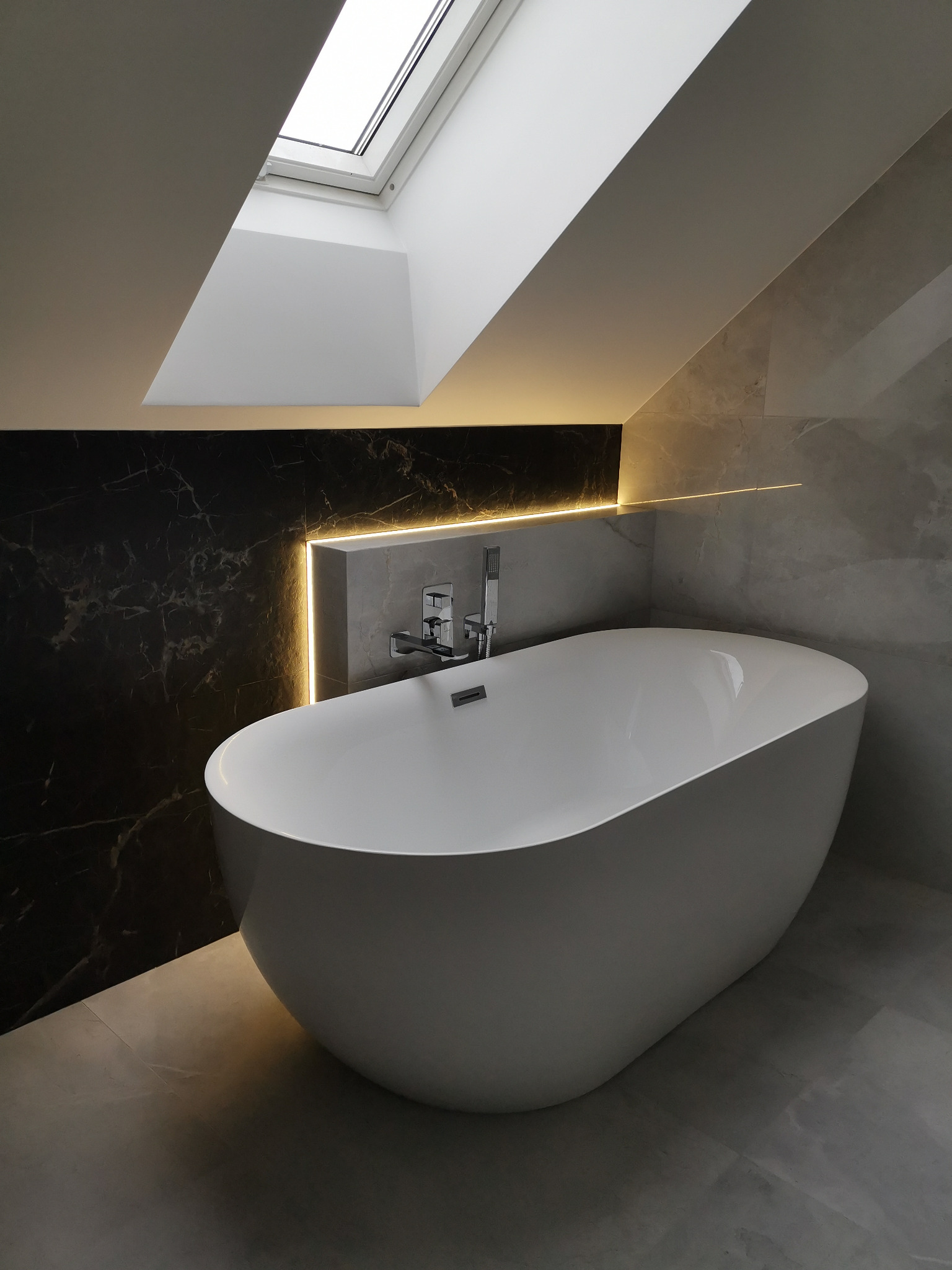 Nowoczesna, wolnostojąca wanna w łazience na poddaszu w Skierniewicach, z oknem dachowym i podświetleniem LED wzdłuż ściany wyłożonej czarnym marmurem.