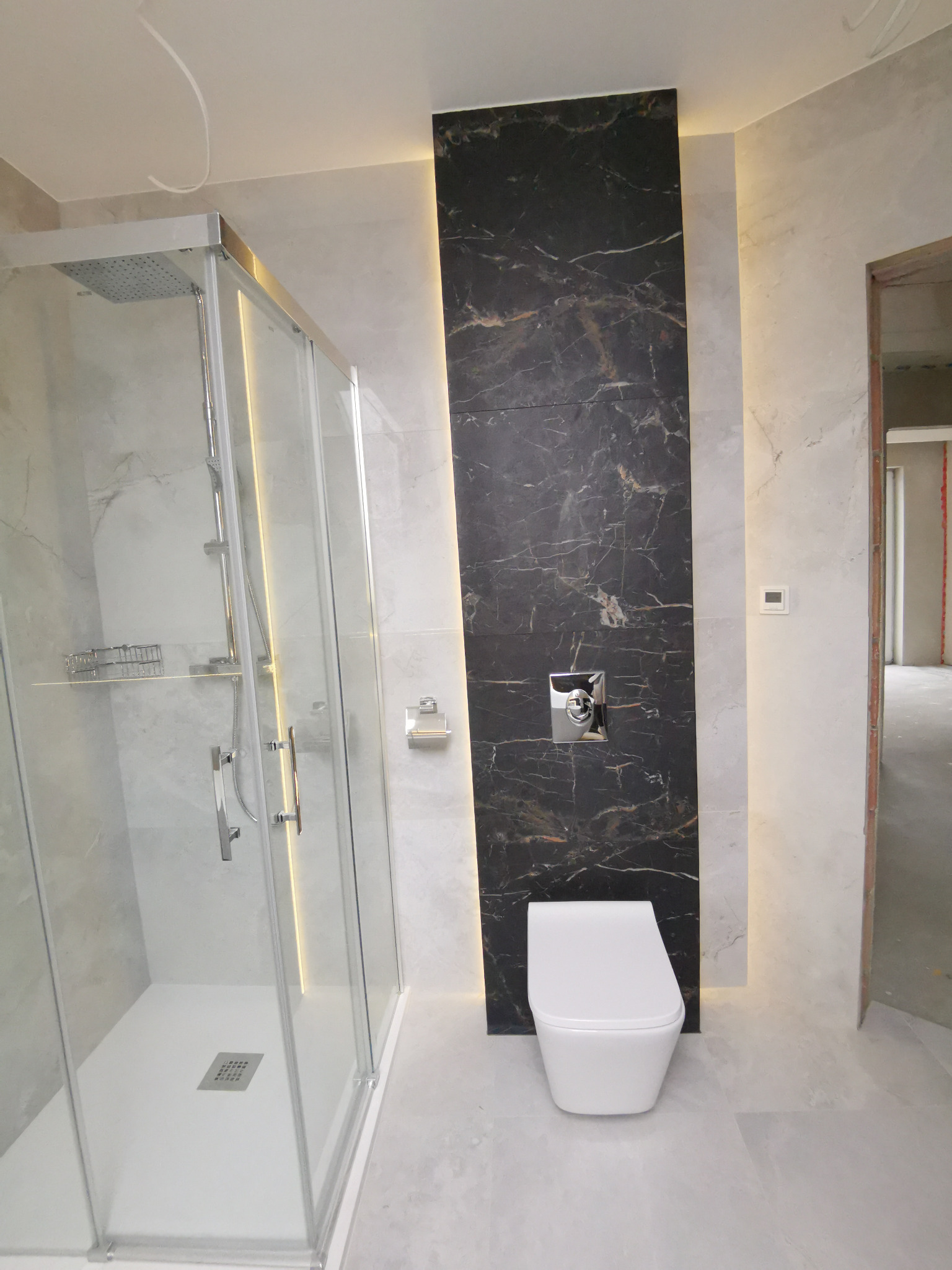 Wykończona łazienka z kabiną prysznicową typu walk-in, wiszącą toaletą i czarną ścianą z marmurowym wzorem podświetloną paskiem LED.