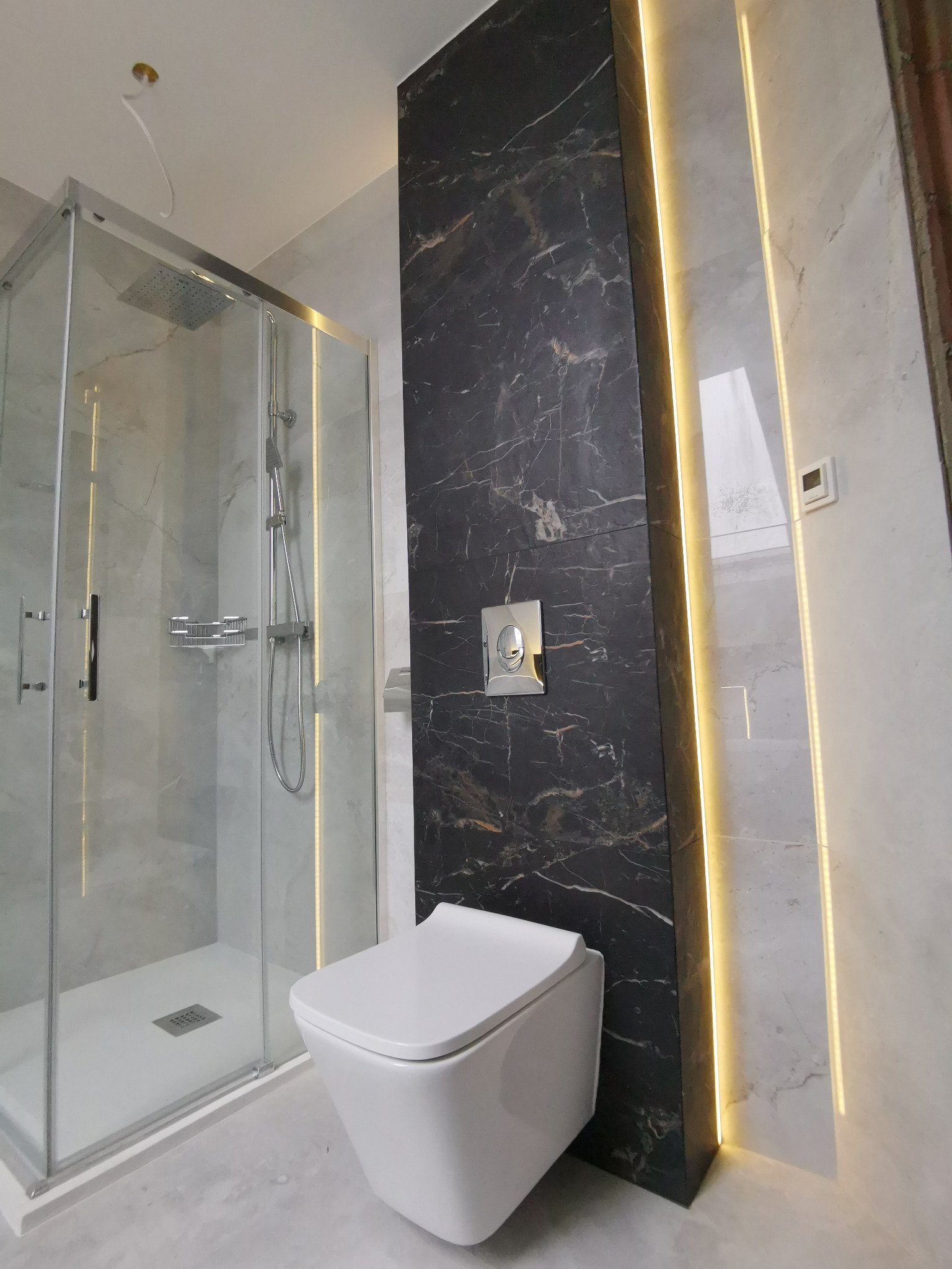 Nowoczesna łazienka z kabiną prysznicową ze srebrnymi ramami i deszczownicą, wisząca toaleta z chromowanym przyciskiem spłukiwania na tle ściany z czarnego marmuru z białymi żyłami, oświetlenie LED...