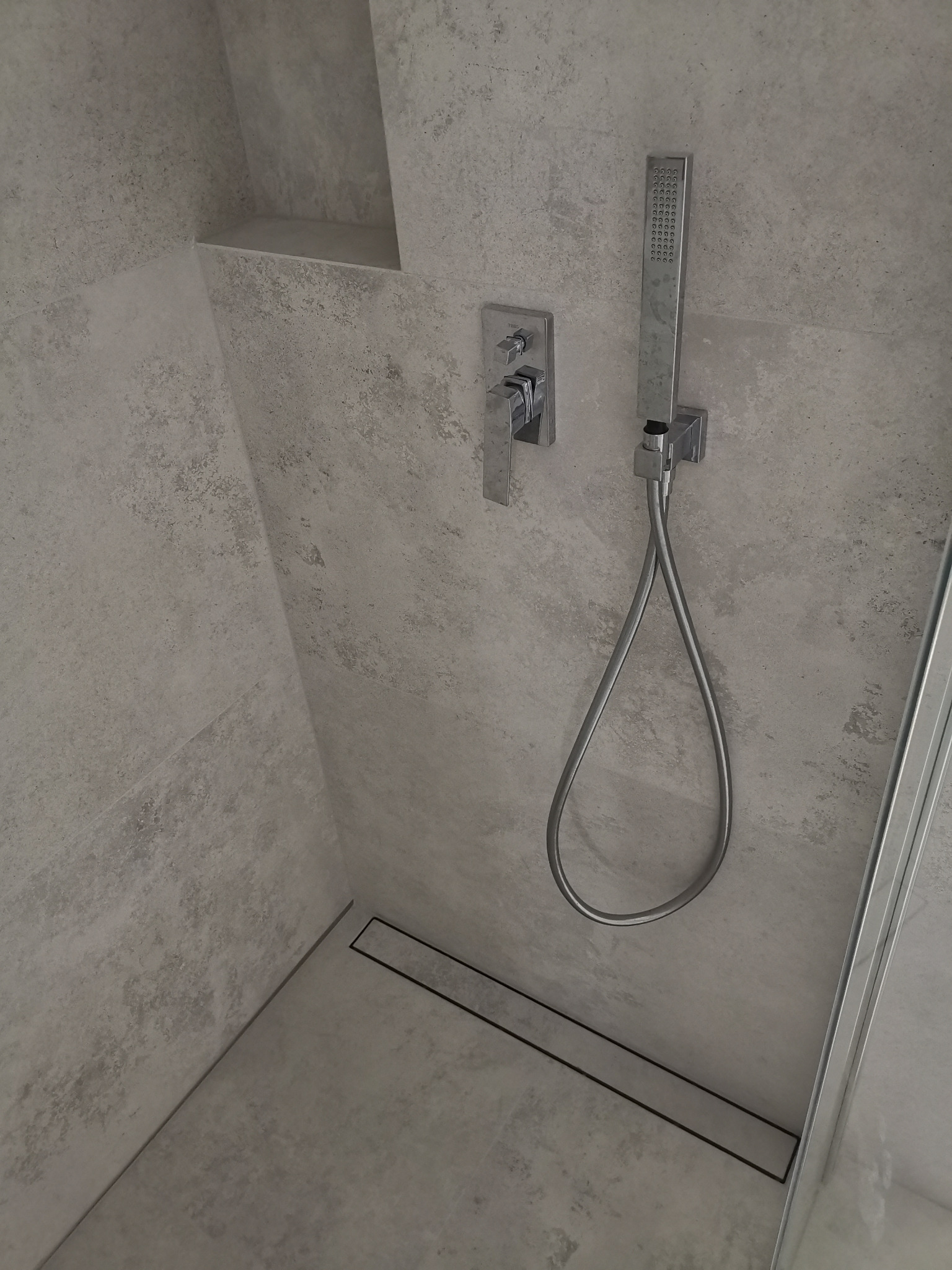 Szczegółowe ujęcie wykończonej kabiny prysznicowej z szarymi płytkami imitującymi beton, metalową armaturą, odpływem liniowym w podłodze i półką w ścianie.