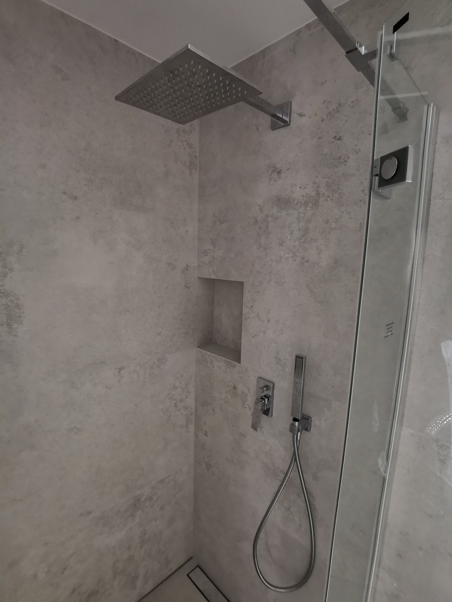 Nowoczesna kabina prysznicowa z deszczownicą, szarymi płytkami imitującymi beton i wnęką na kosmetyki, widoczna szklana ściana kabiny.