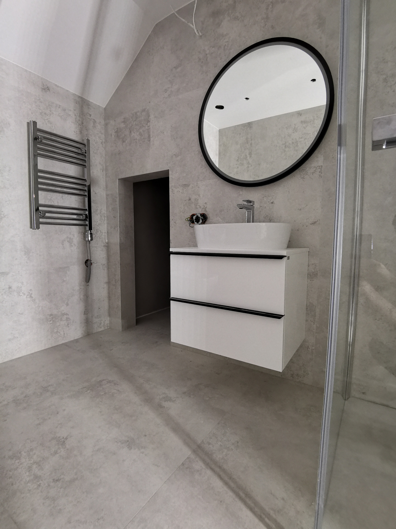 Nowoczesna łazienka w szarościach z okrągłym lustrem, grzejnikiem drabinkowym i białą szafką pod umywalką, widoczna kabina prysznicowa z przezroczystego szkła.