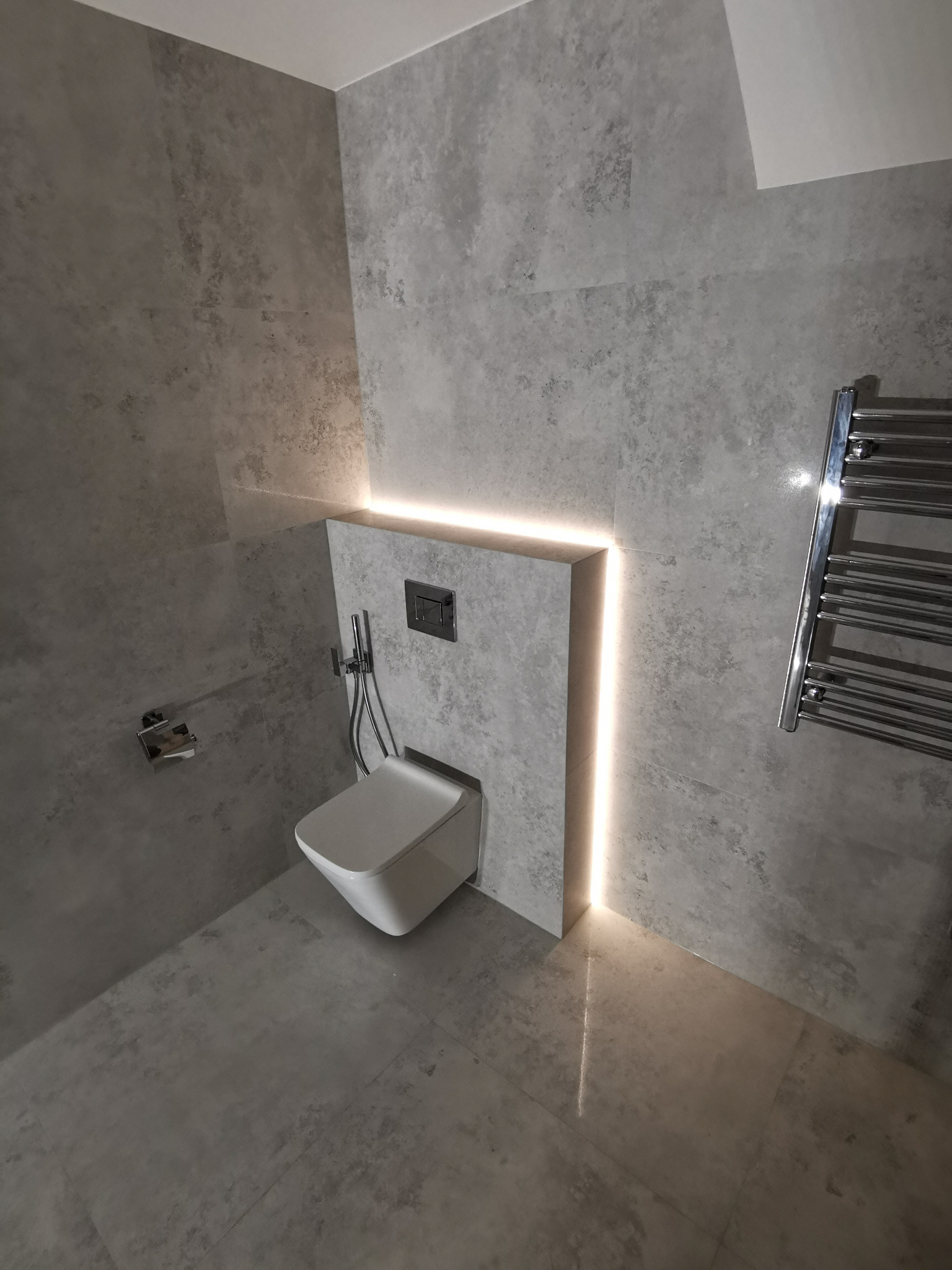 Nowoczesna łazienka w odcieniach szarości z podświetlaną wnęką za toaletą podwieszaną i grzejnikiem drabinkowym.