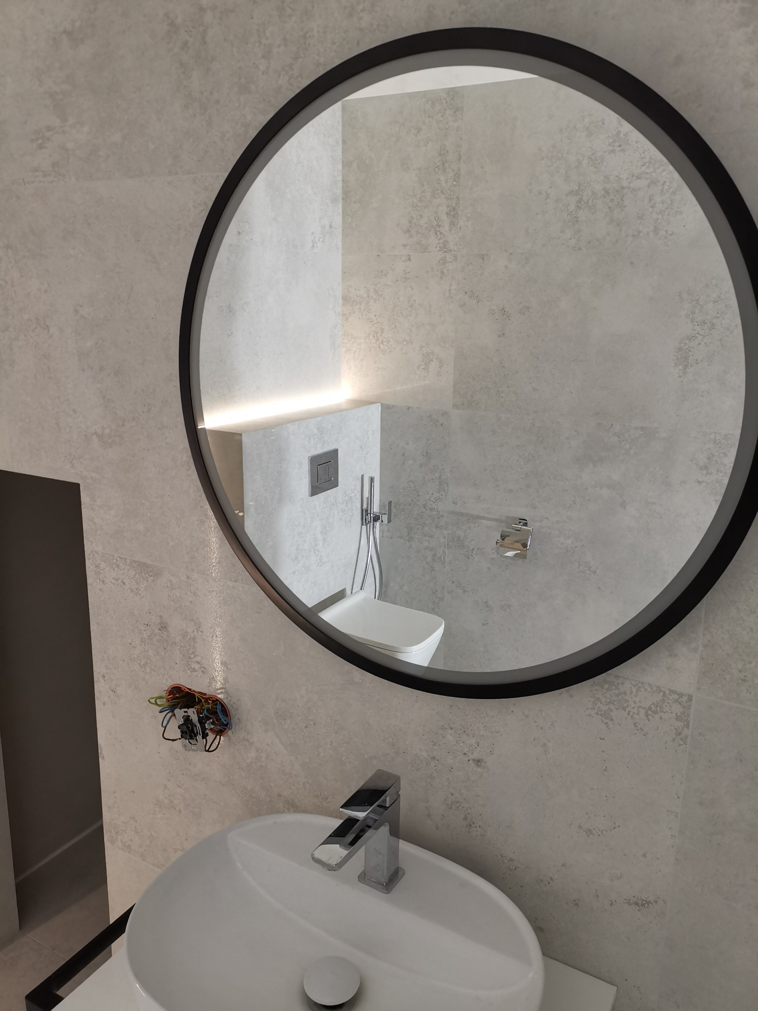 Okrągłe lustro z czarną ramą odbija fragment łazienki z podświetleniem LED, widoczna umywalka z baterią, w tle toaleta, na ścianie widoczne kable wystające ze ściany przed montażem oświetlenia.