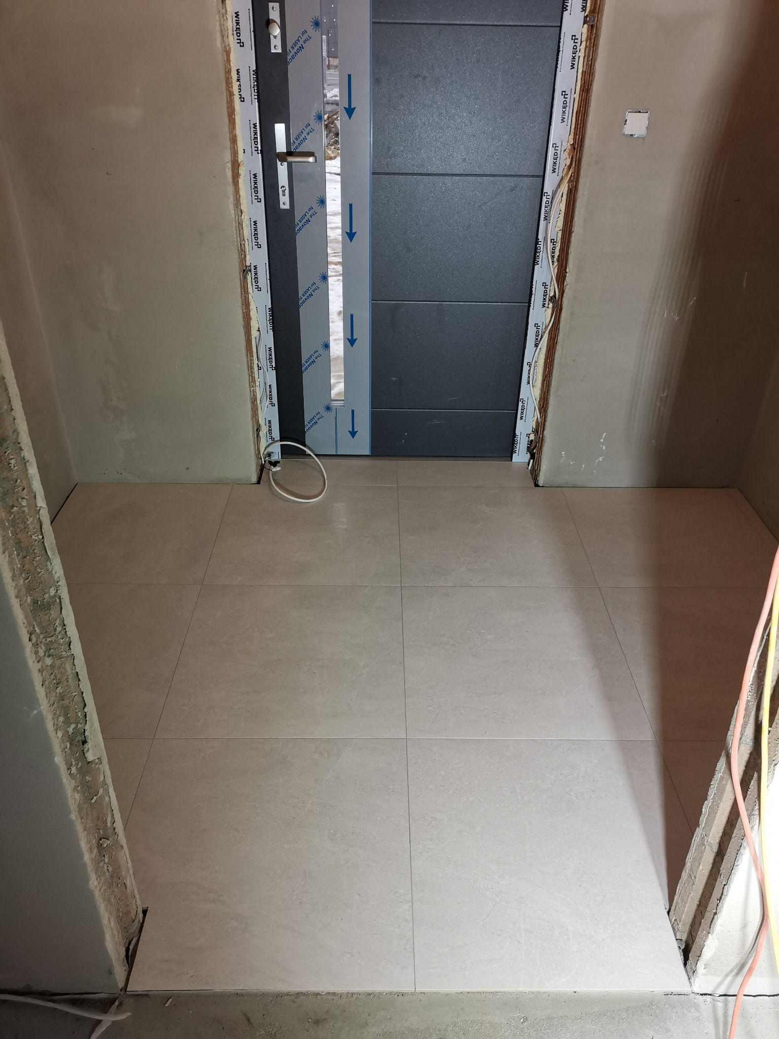 Świeżo ułożona, jasna podłoga z płytek w przedpokoju, widoczne ściany w trakcie prac wykończeniowych, drzwi wejściowe z folią ochronną na ościeżnicy.