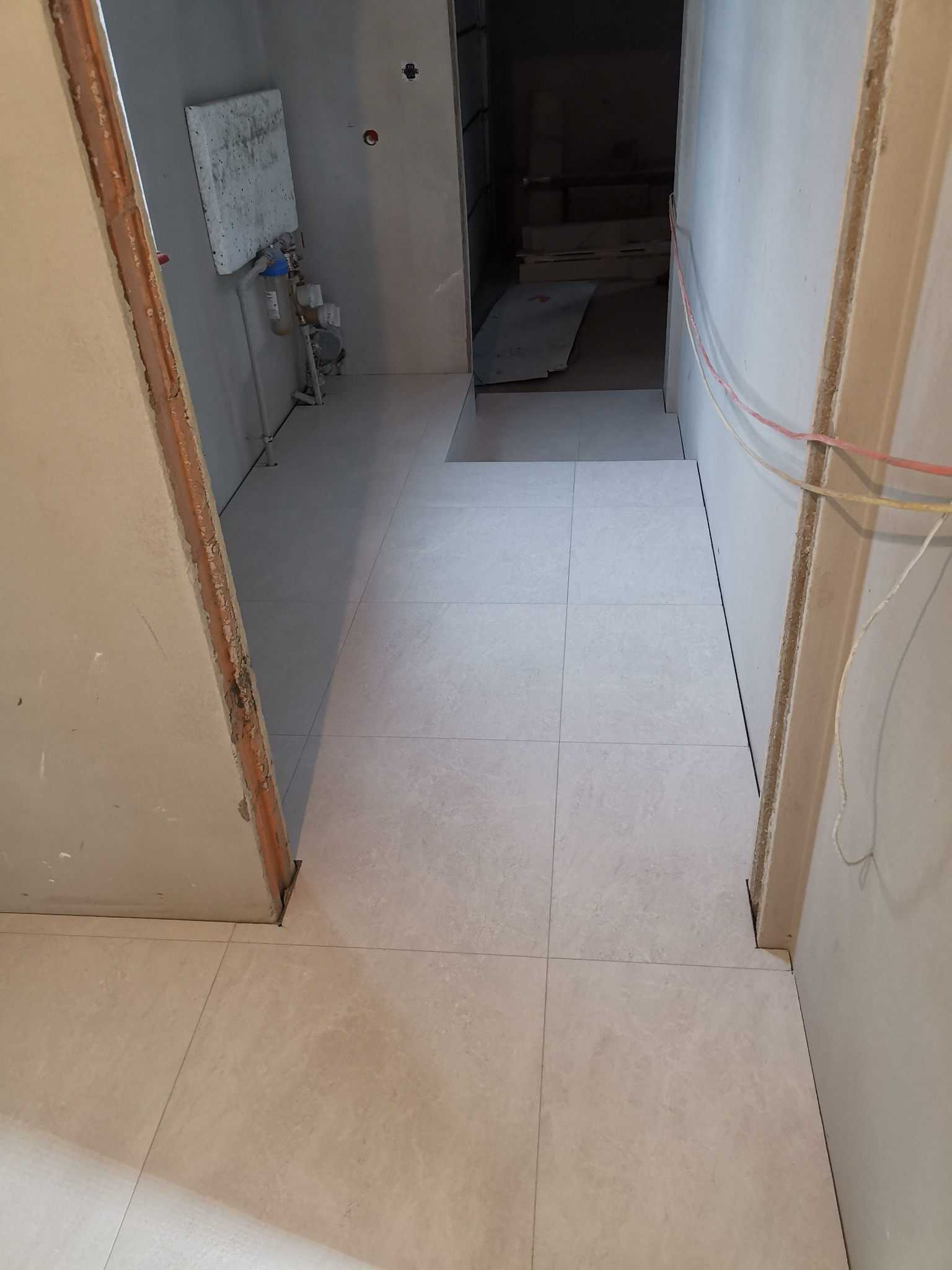 Ułożone płytki podłogowe w jasnym odcieniu szarości, widok z progu drzwi, ściany przygotowane do malowania, instalacja wodna widoczna w tle.