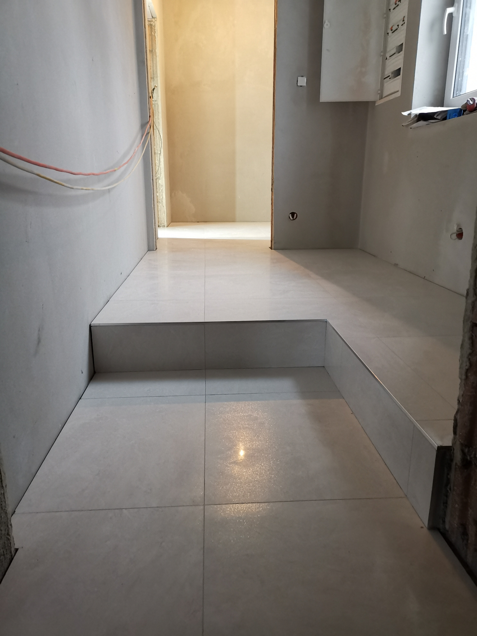 Świeżo ułożona, jasna, kwadratowa podłoga z płytek w łazience na dwóch poziomach, widoczne niezabezpieczone kable elektryczne na ścianie, oraz skrzynka elektryczna obok okna.