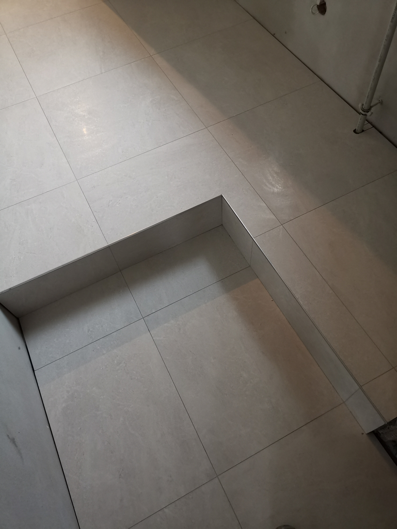 Ułożone jasnoszare płytki podłogowe w łazience, widoczny fragment obudowy wanny z metalowym wykończeniem krawędzi, rura instalacyjna przy ścianie.