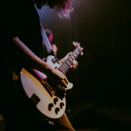 Gitarzysta grający na białej gitarze elektrycznej podczas koncertu, z klawiszowcem w tle, oświetlony kolorowymi światłami scenicznymi.