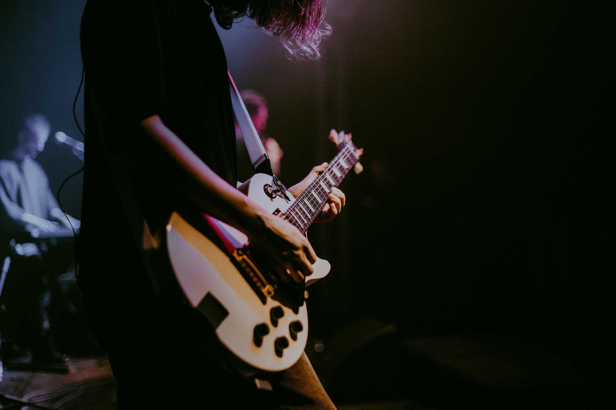 Gitarzysta grający na białej gitarze elektrycznej podczas koncertu, z klawiszowcem w tle, oświetlony kolorowymi światłami scenicznymi.