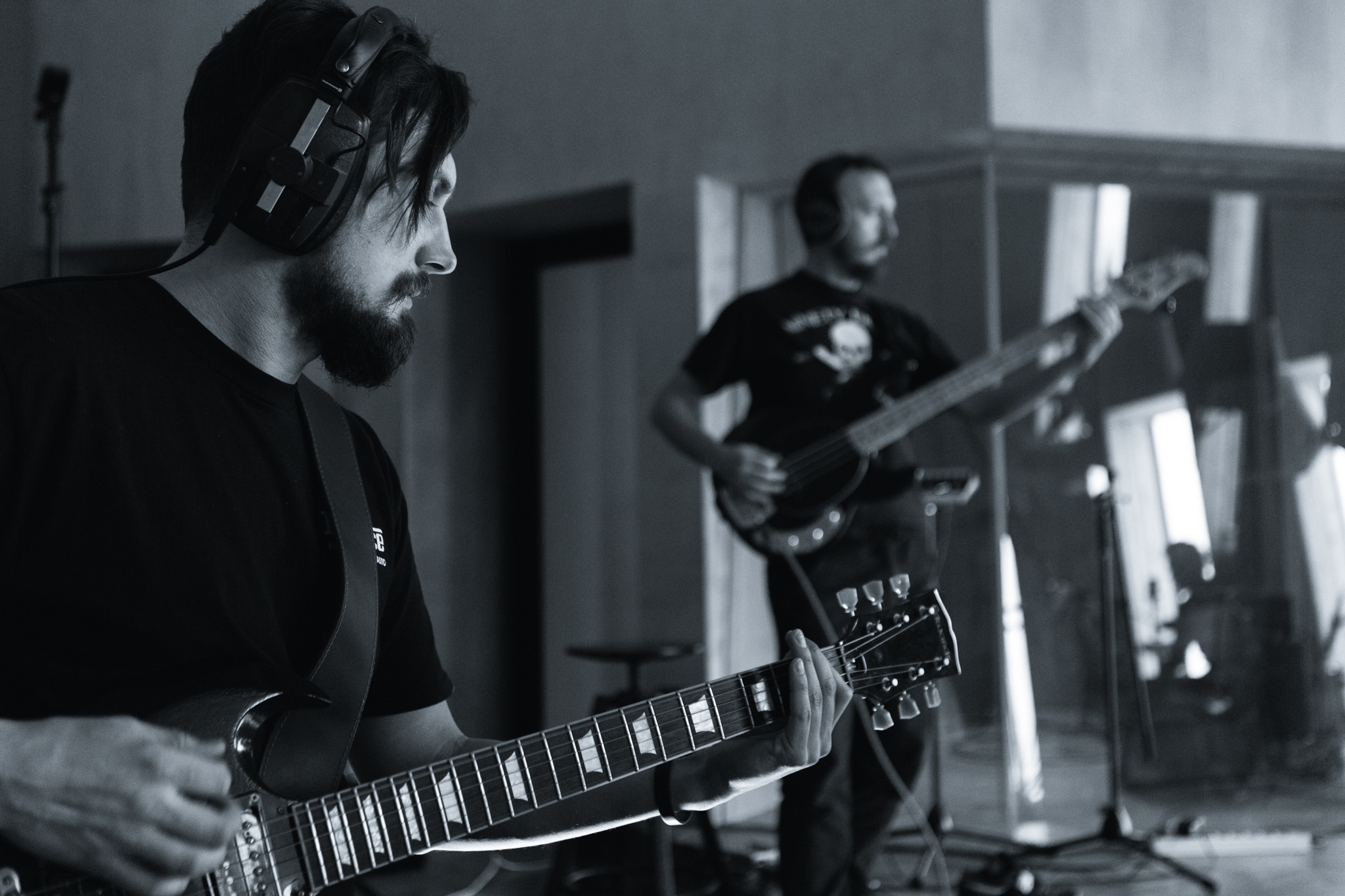 Monochromatyczne ujęcie dwóch muzyków w studio: pierwszy plan - gitarzysta z brodą i słuchawkami na uszach grający na gitarze elektrycznej, drugi plan - basista z gitarą basową i słuchawkami...