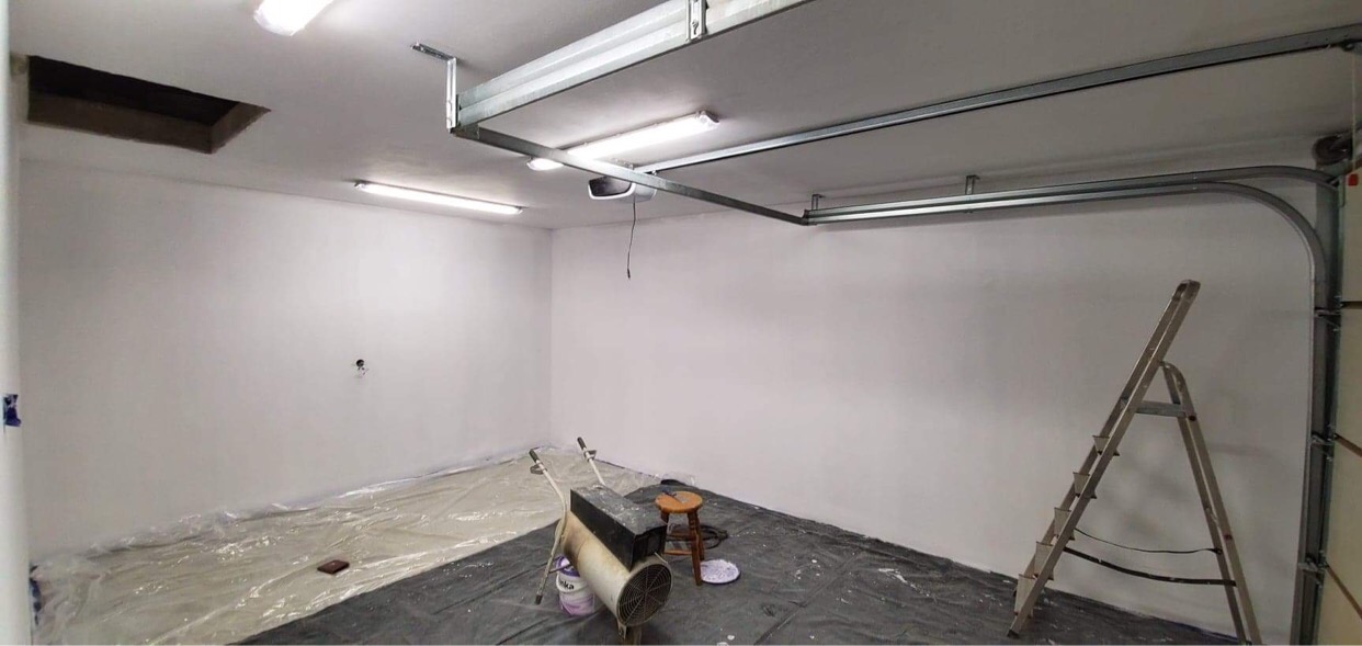 Wnętrze garażu po malowaniu na biało, widoczne zabezpieczone folią podłogi, drabina oparta o ścianę, nagrzewnica i taboret malarski.