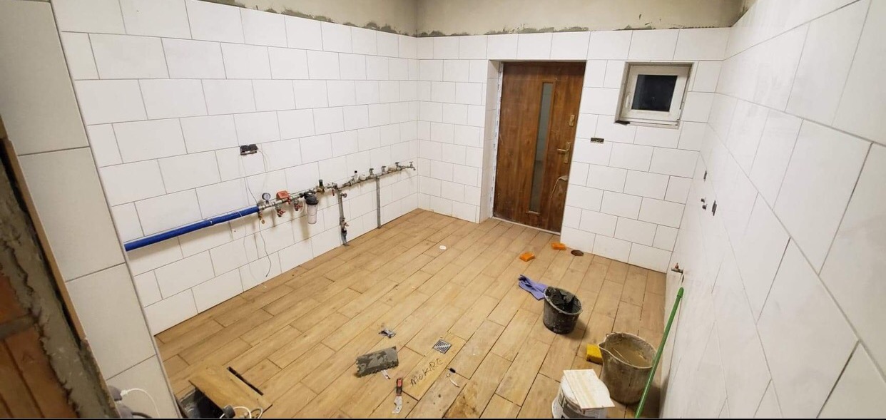 Wnętrze łazienki w trakcie remontu: białe, prostokątne płytki na ścianach, podłoga wyłożona płytkami imitującymi drewno, widoczne instalacje wodne, narzędzia i materiały budowlane, częściowo...