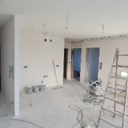 Wnętrze w trakcie prac przygotowawczych do malowania: ściany po szpachlowaniu, widoczne puszki z farbą, drabina z lampą roboczą, instalacja elektryczna wyprowadzona ze ścian i sufitu.