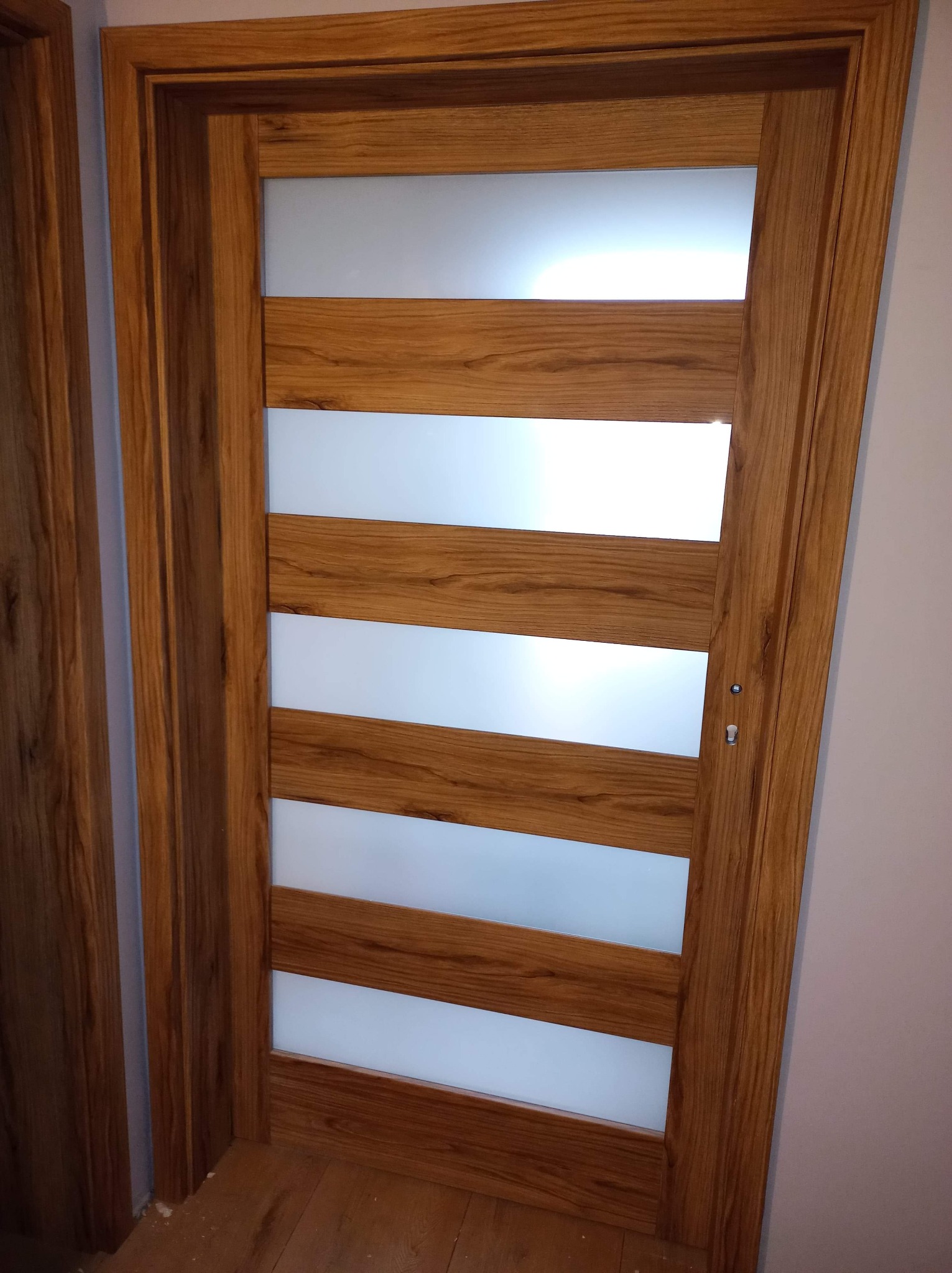 Drewniane drzwi wewnętrzne z czterema poziomymi, matowymi przeszkleniami, widoczne z bliska, z delikatnym usłojeniem drewna i srebrną klamką.