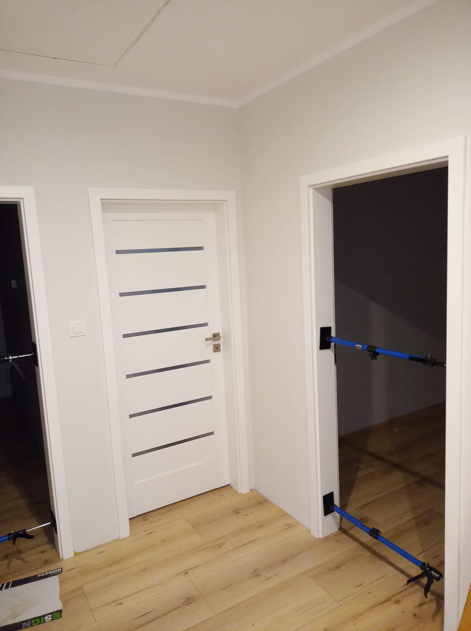 Wnętrze mieszkania po remoncie: białe ściany, drewnopodobna podłoga, białe drzwi z poziomymi przeszkleniami, zabezpieczenia na ościeżnicach, widoczne narzędzia.