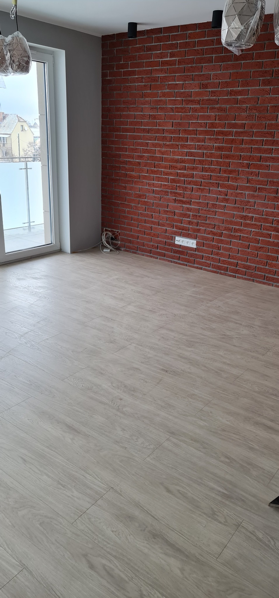 Puste pomieszczenie z panelami podłogowymi w odcieniach szarości, ściana z czerwonej cegły, okno balkonowe i widoczne przewody elektryczne przy podłodze.