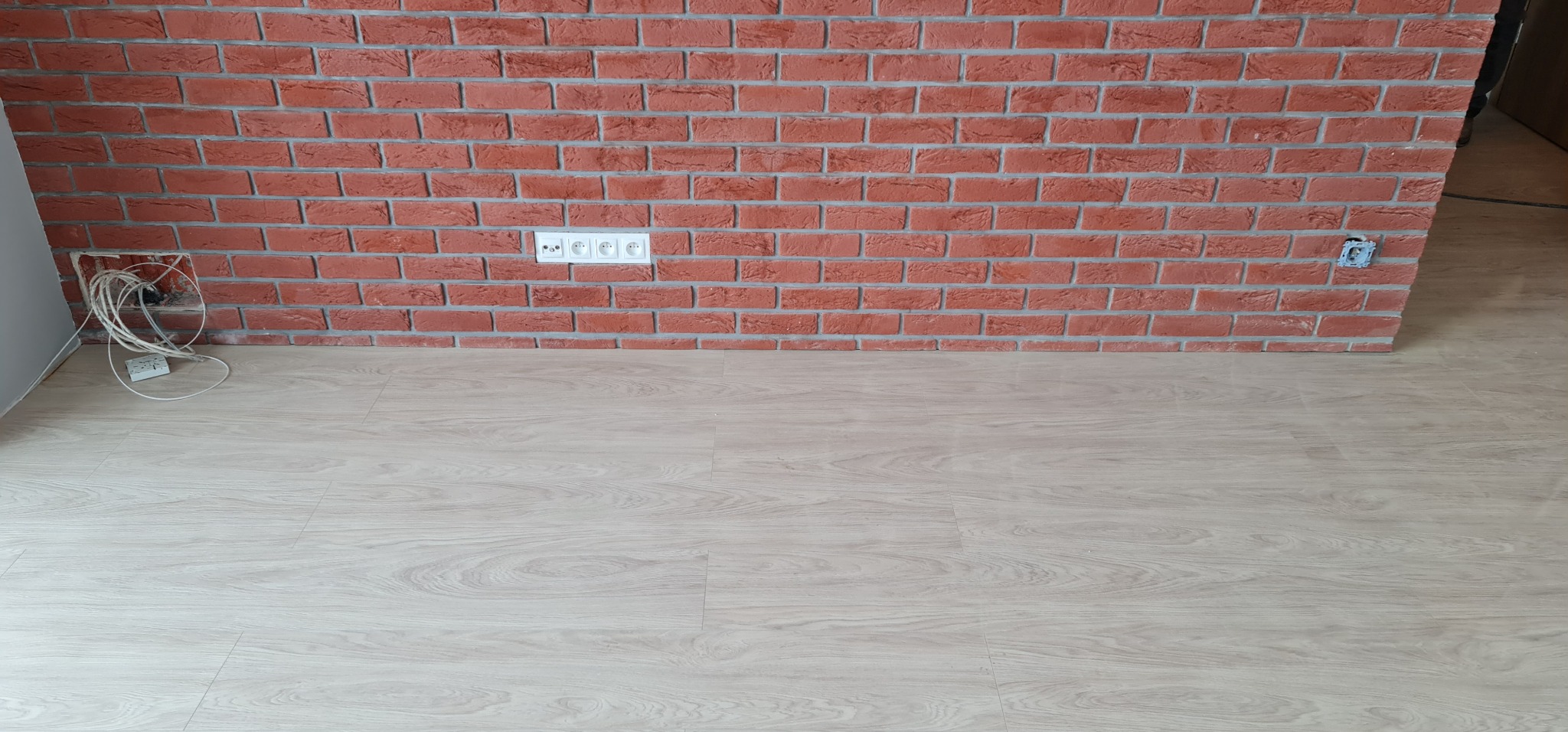 Wykończona ściana z cegły klinkierowej z gniazdkami elektrycznymi na jasnej podłodze z paneli, widoczne kable wystające z otworu w ścianie po lewej stronie.
