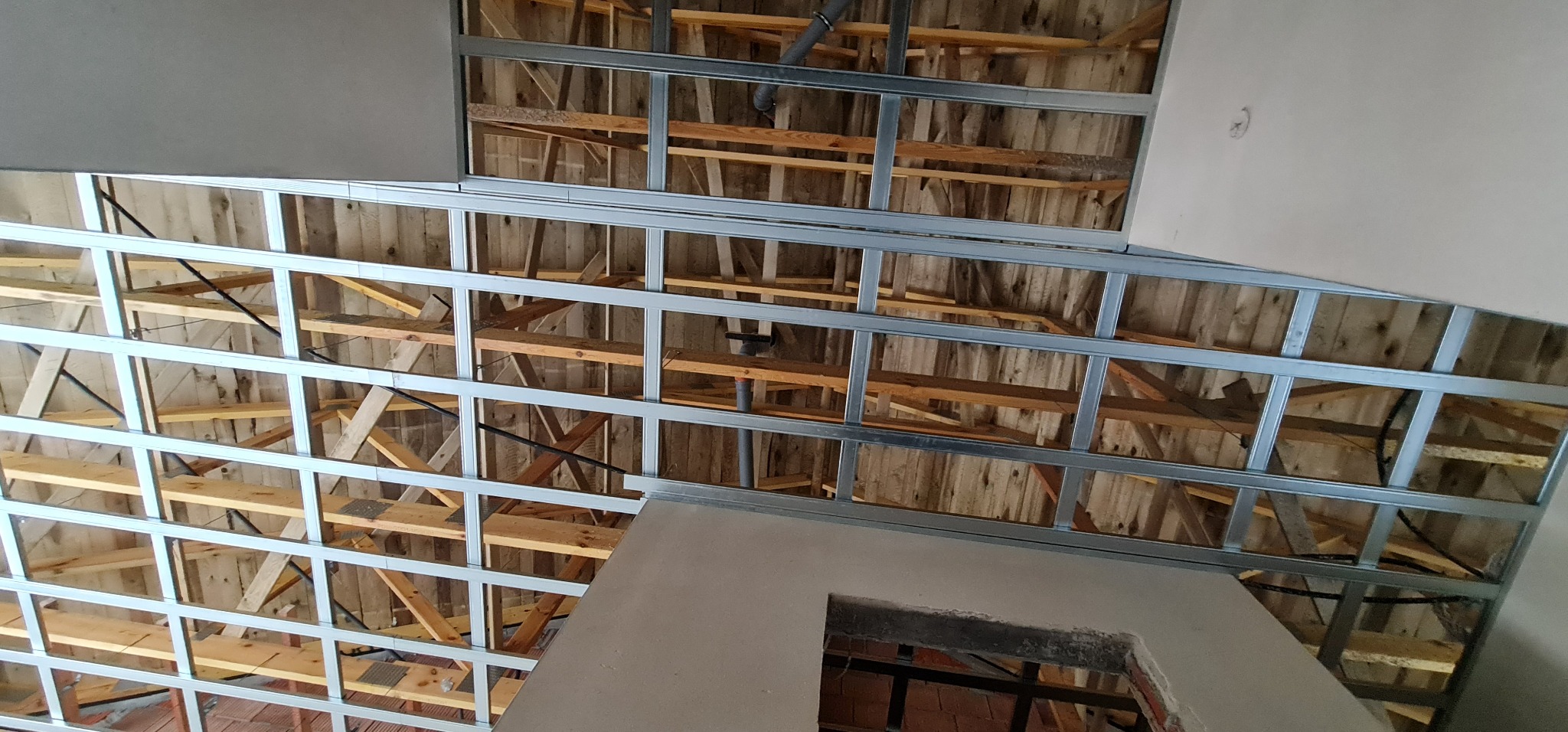 Konstrukcja metalowa sufitu podwieszanego zamontowana do drewnianej więźby dachowej, widoczne rury instalacyjne i fragment ściany z otworem.
