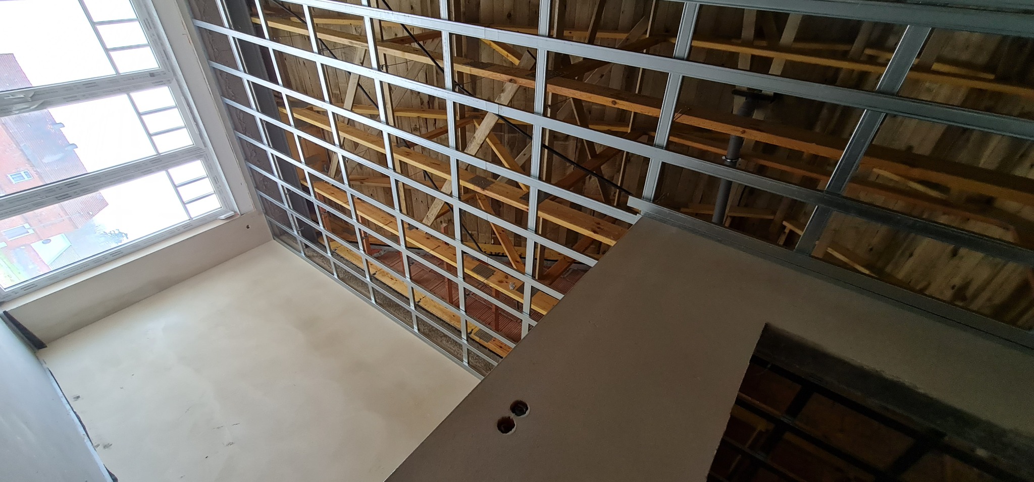 Stelaż sufitu podwieszanego z profili metalowych, widoczne drewniane elementy konstrukcyjne dachu, okno z widokiem na budynek z czerwonej cegły