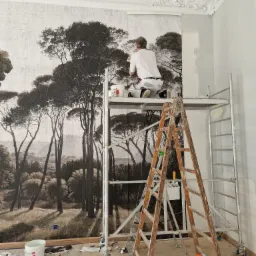 Pracownik na rusztowaniu w Olsztynie aplikuje tapetę z motywem leśnym na ścianę w pomieszczeniu z ozdobnym sufitem, widoczne narzędzia i resztki tapety na podłodze.