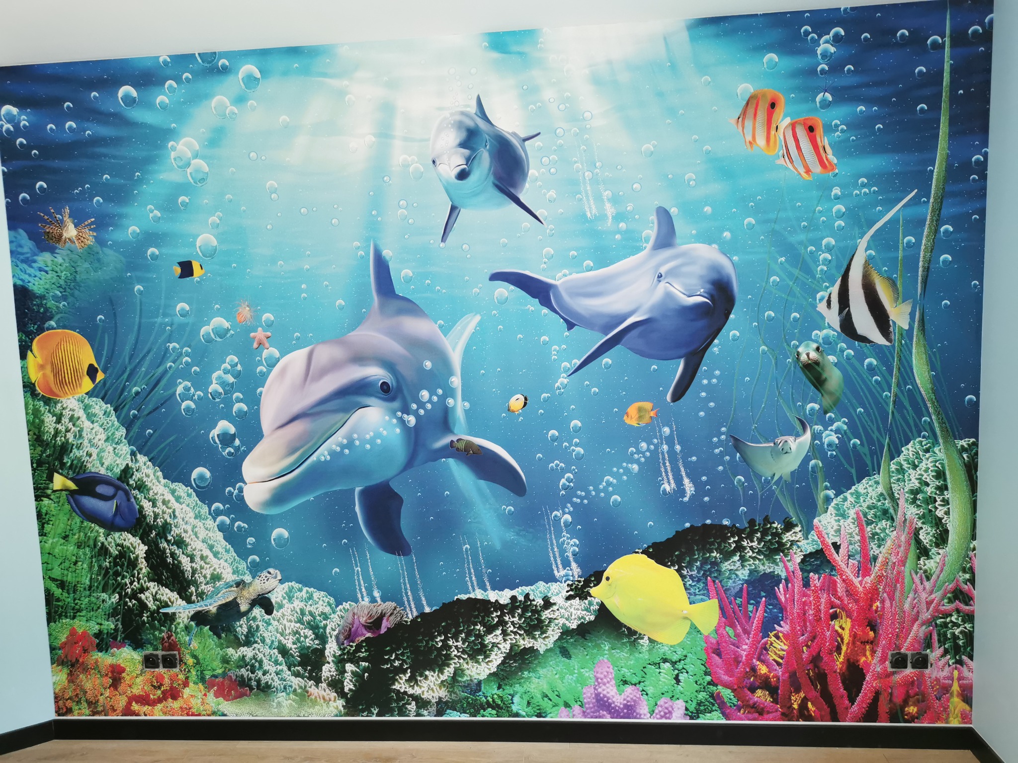 Ściana pokryta fototapetą z motywem podwodnego świata: delfiny, ryby tropikalne, koralowce i bąbelki powietrza w błękitnej wodzie, imitacja akwarium.