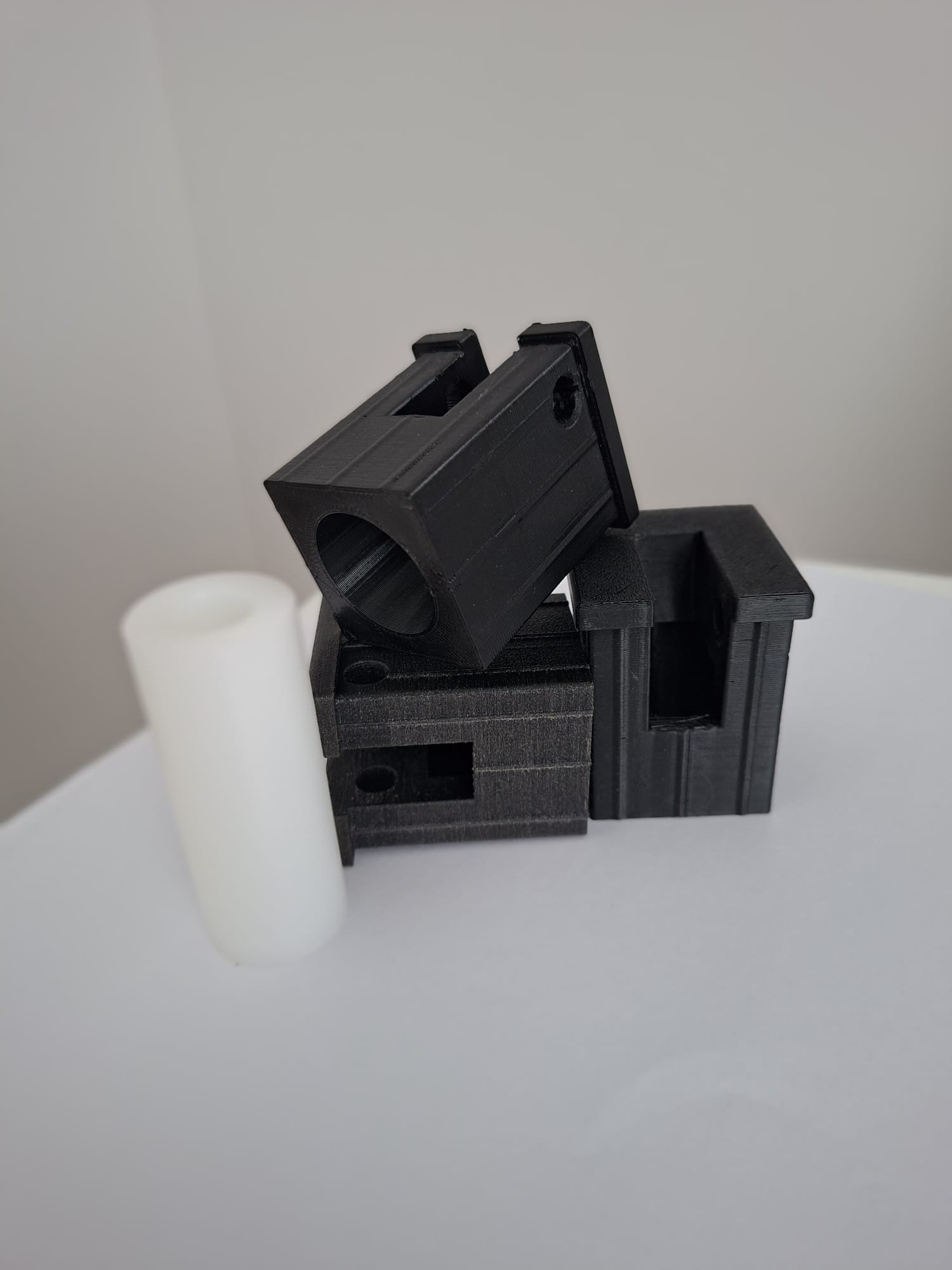 Czarne, modułowe elementy z widoczną strukturą druku 3D, ułożone obok białego walca na jasnym tle. Detal precyzyjnego wykonania.