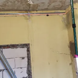 Wnętrze w trakcie prac remontowych: ściana z żółtą farbą, otwór w ścianie z widocznymi bloczkami, drabina, poziomica laserowa wyznaczająca linię na ścianie, sufit z odpadającą farbą.