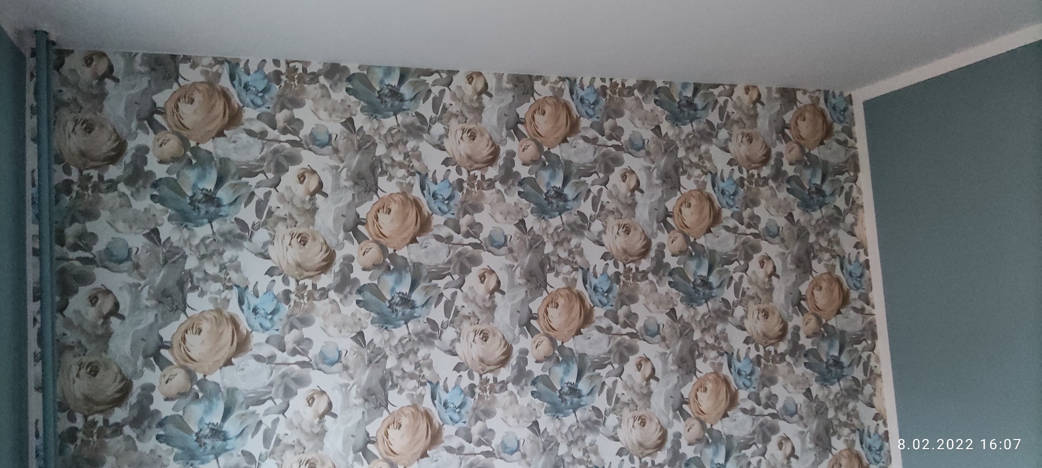 Ściana pokryta tapetą w kwiaty w odcieniach beżu i błękitu, fragment ściany pomalowanej na jednolity błękitny kolor oraz rura w tym samym kolorze w rogu pomieszczenia.