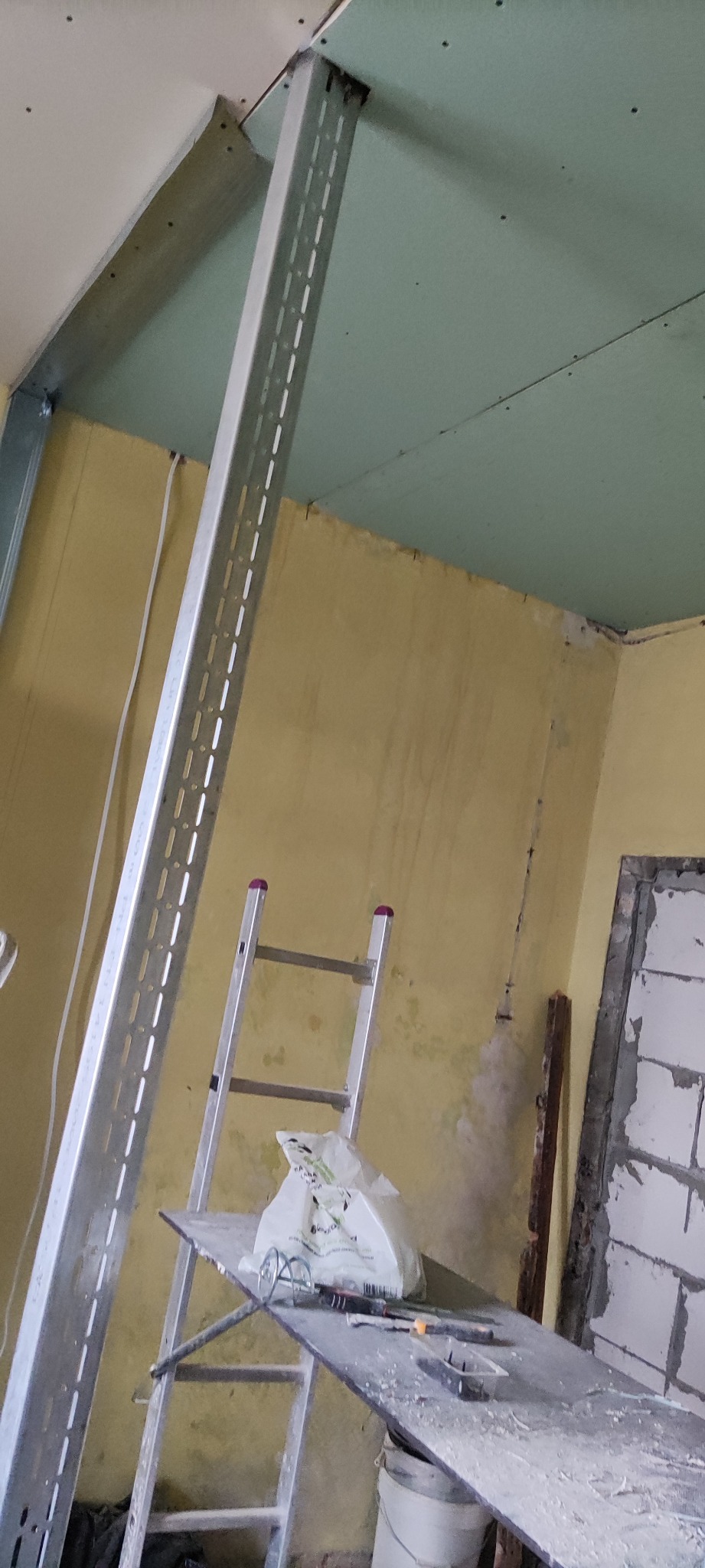 Montaż stelaża z profili stalowych pod płytę gipsowo-kartonową na suficie, widoczna drabina z narzędziami, ściana w trakcie remontu.